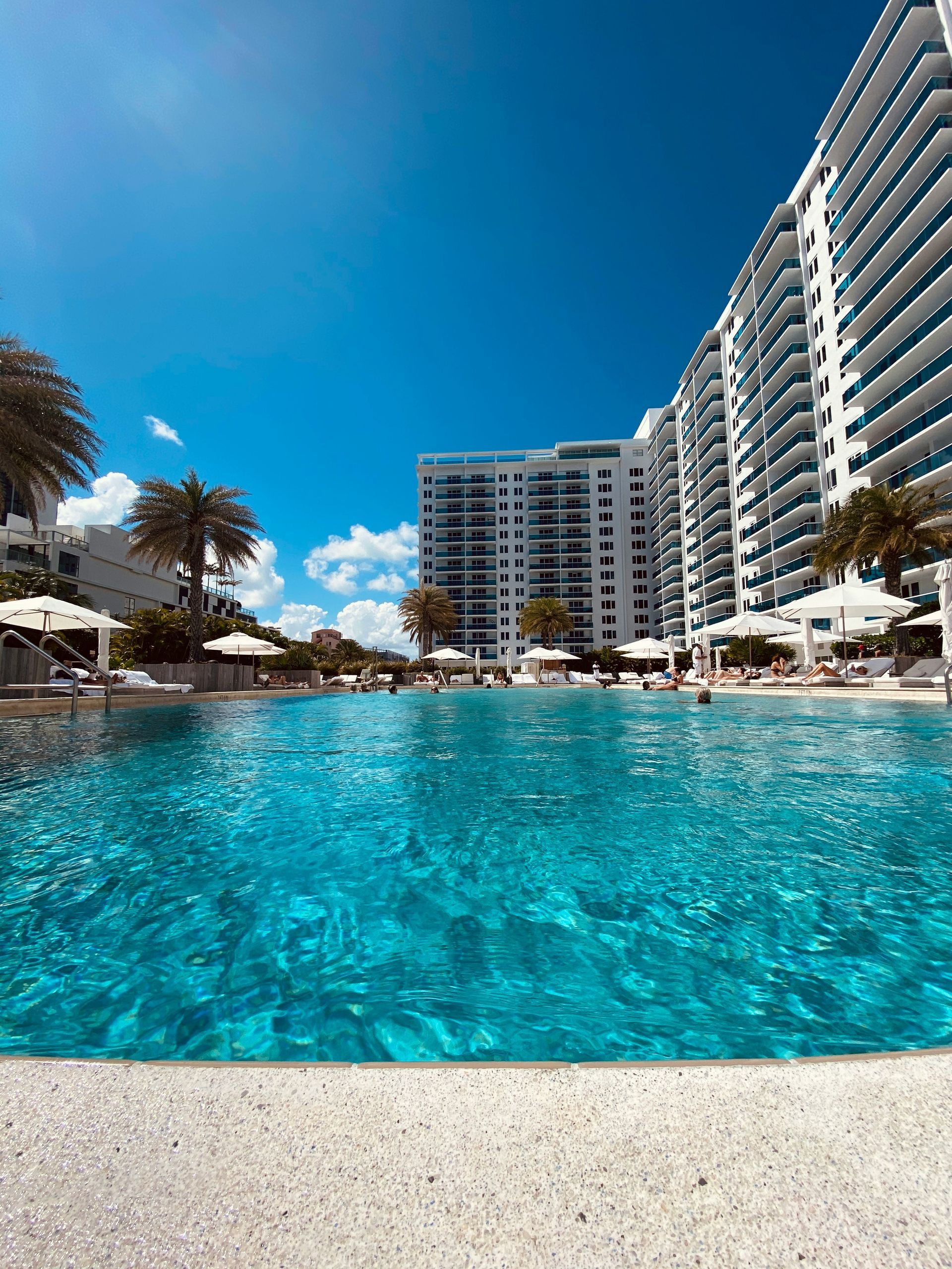 Swimming pool with blue water, white buildings, and bright blue sky. Palm trees.