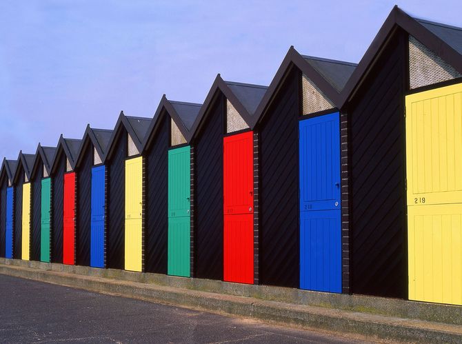 Row of colorful beach huts with black exteriors and brightly colored doors against a clear blue sky.