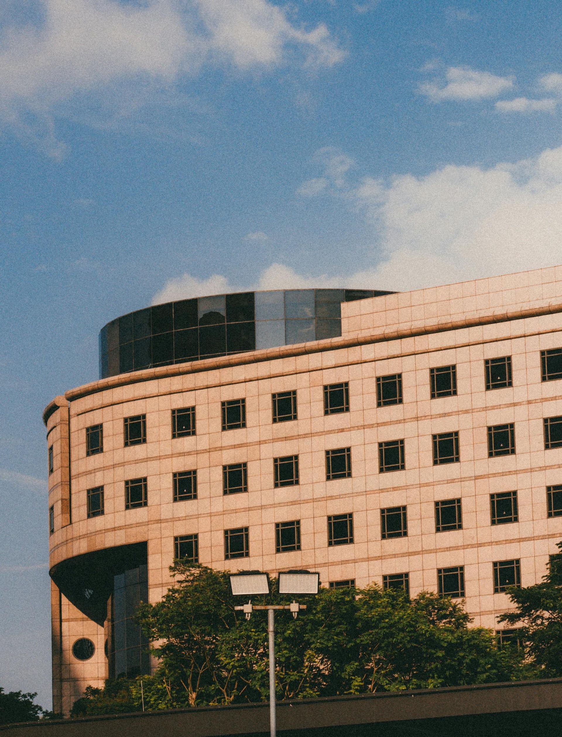 Curved beige building with dark windows, topped by a dark, reflective structure. Blue sky with white clouds.