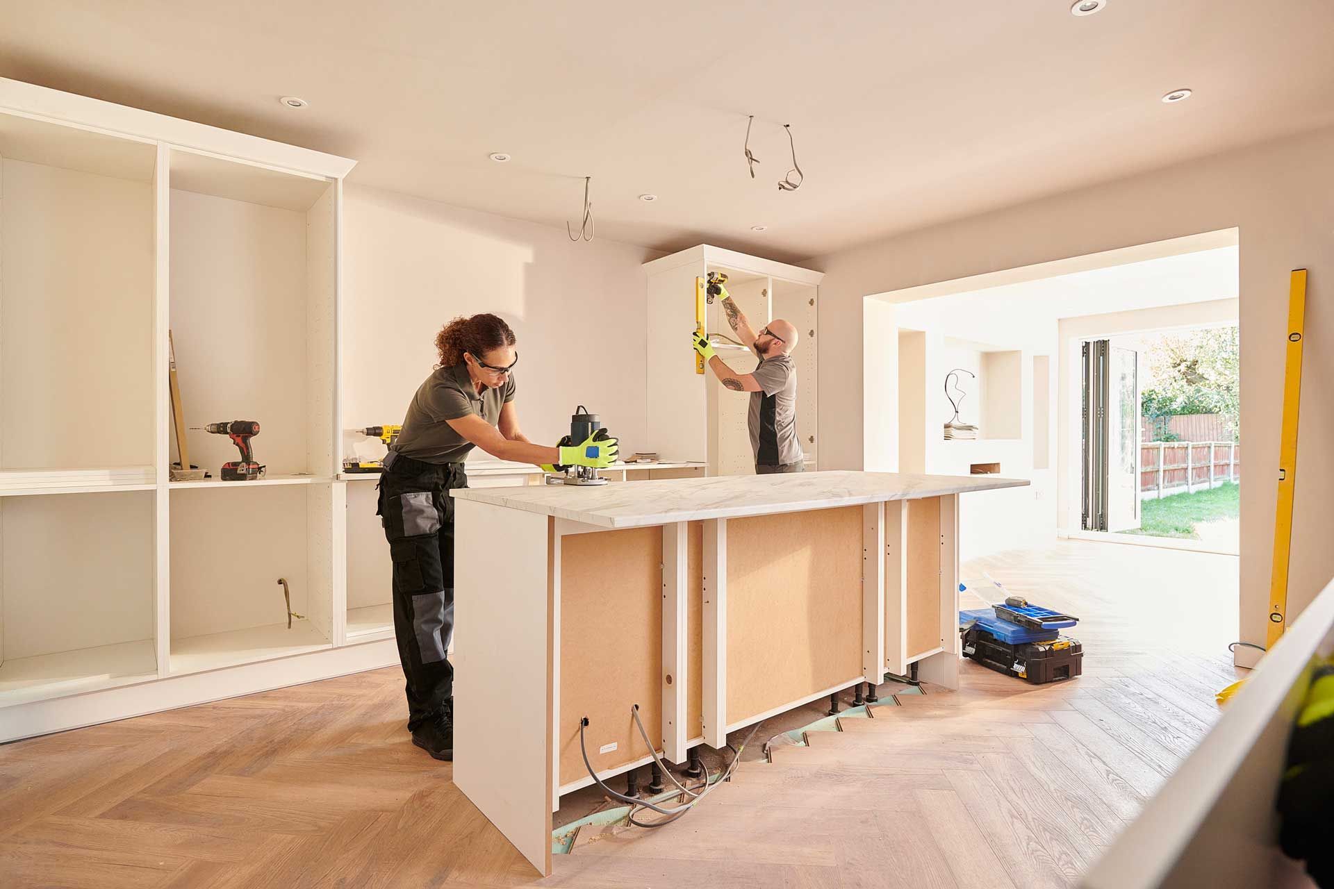 Two people installing kitchen cabinets in a room with hardwood floors.