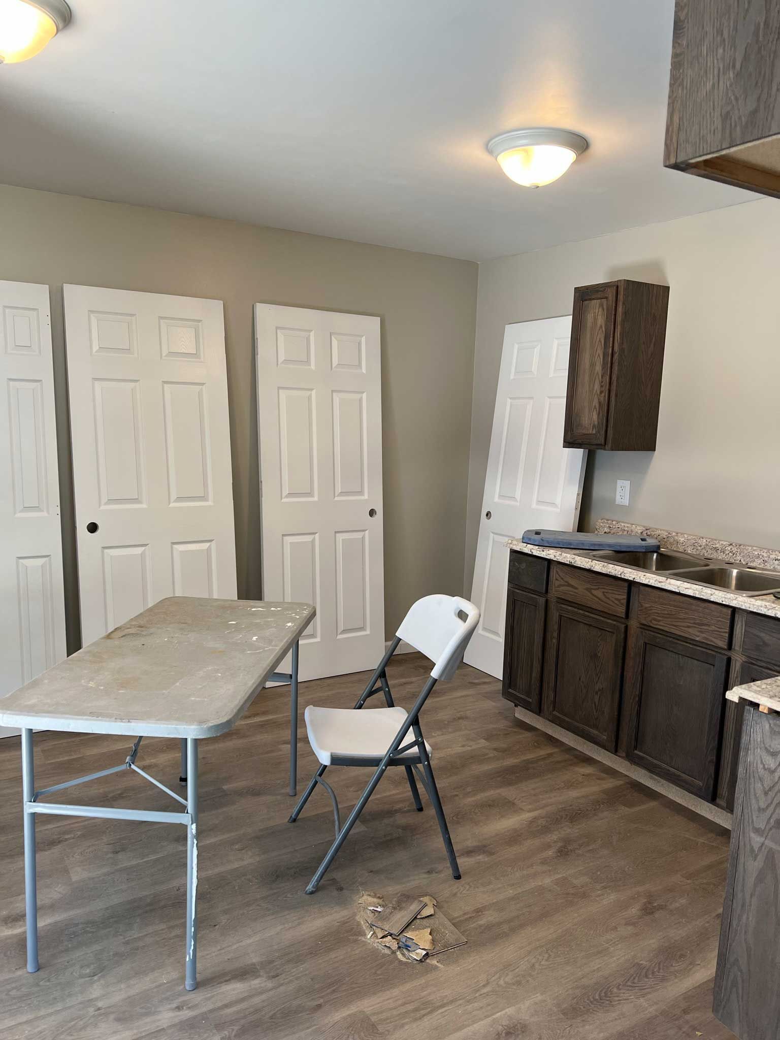 A kitchen renovation in progress, with doors leaning against the wall, a table, chair, and cabinets.