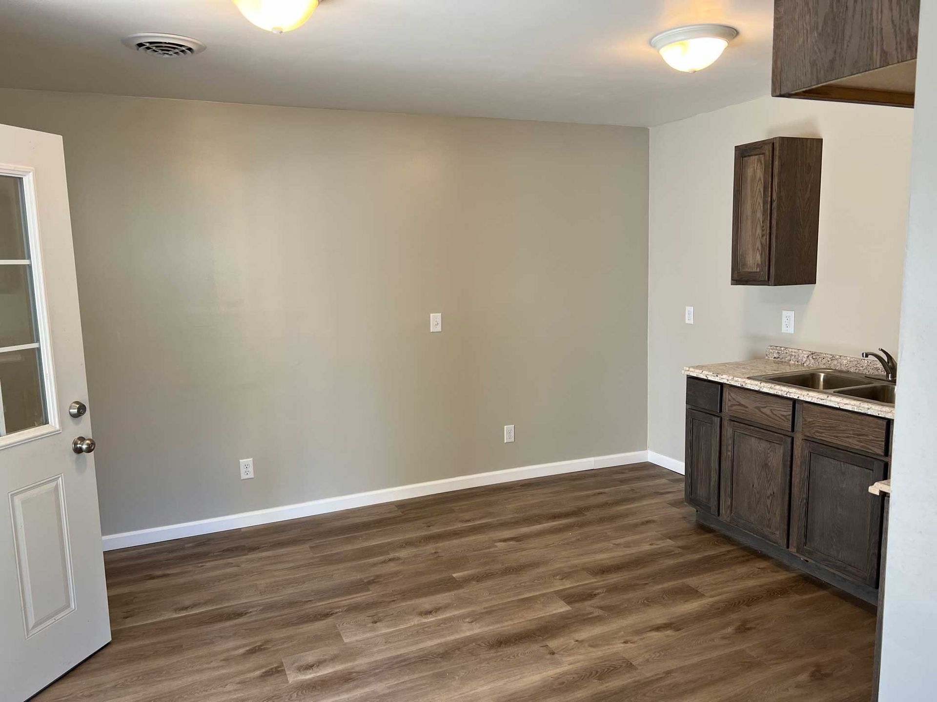 Room interior with neutral walls, wood-look flooring, and a small kitchen area with dark cabinetry.