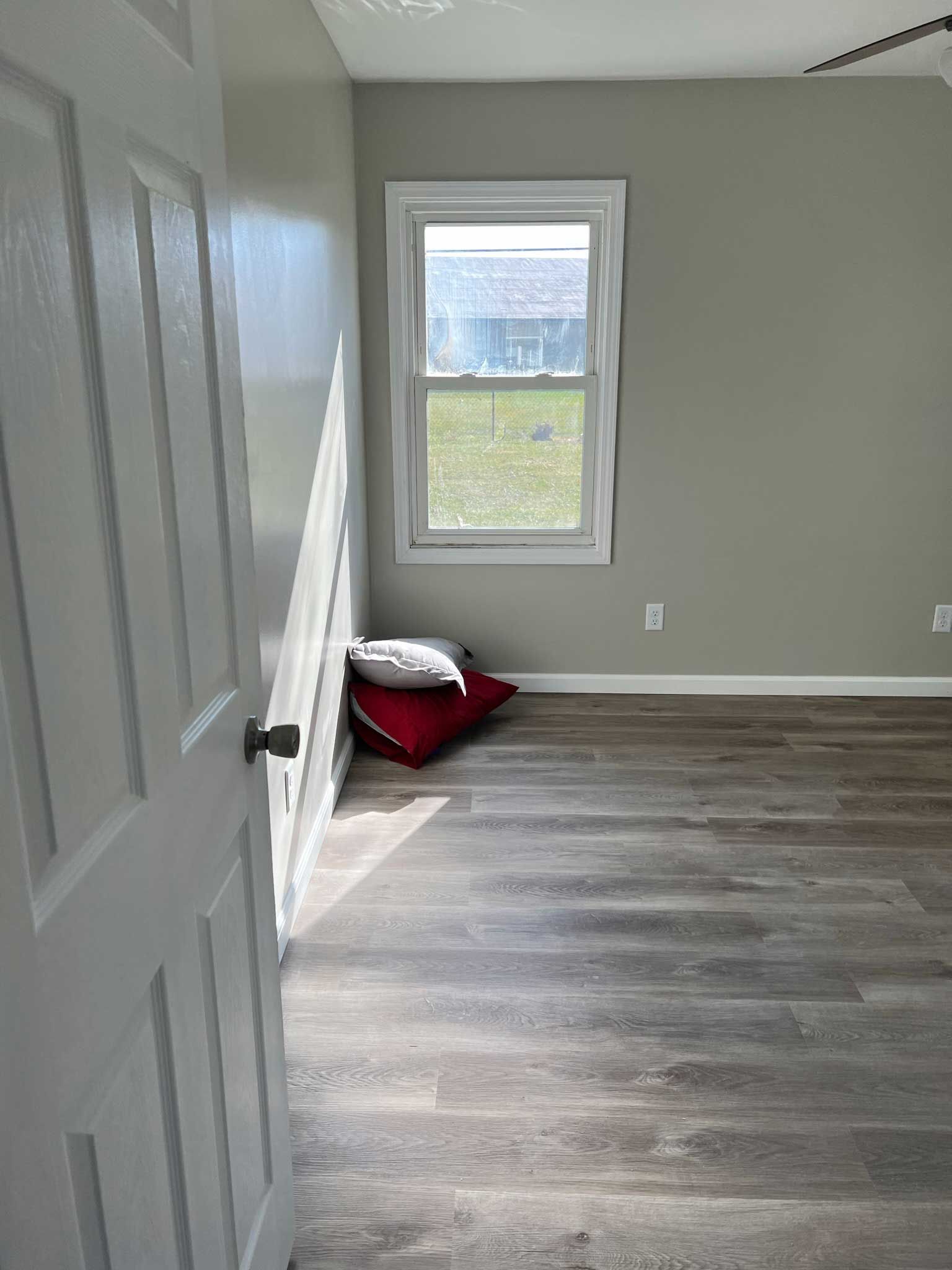 Bedroom with grey walls, white trim, wood-look floor, window, and two pillows.