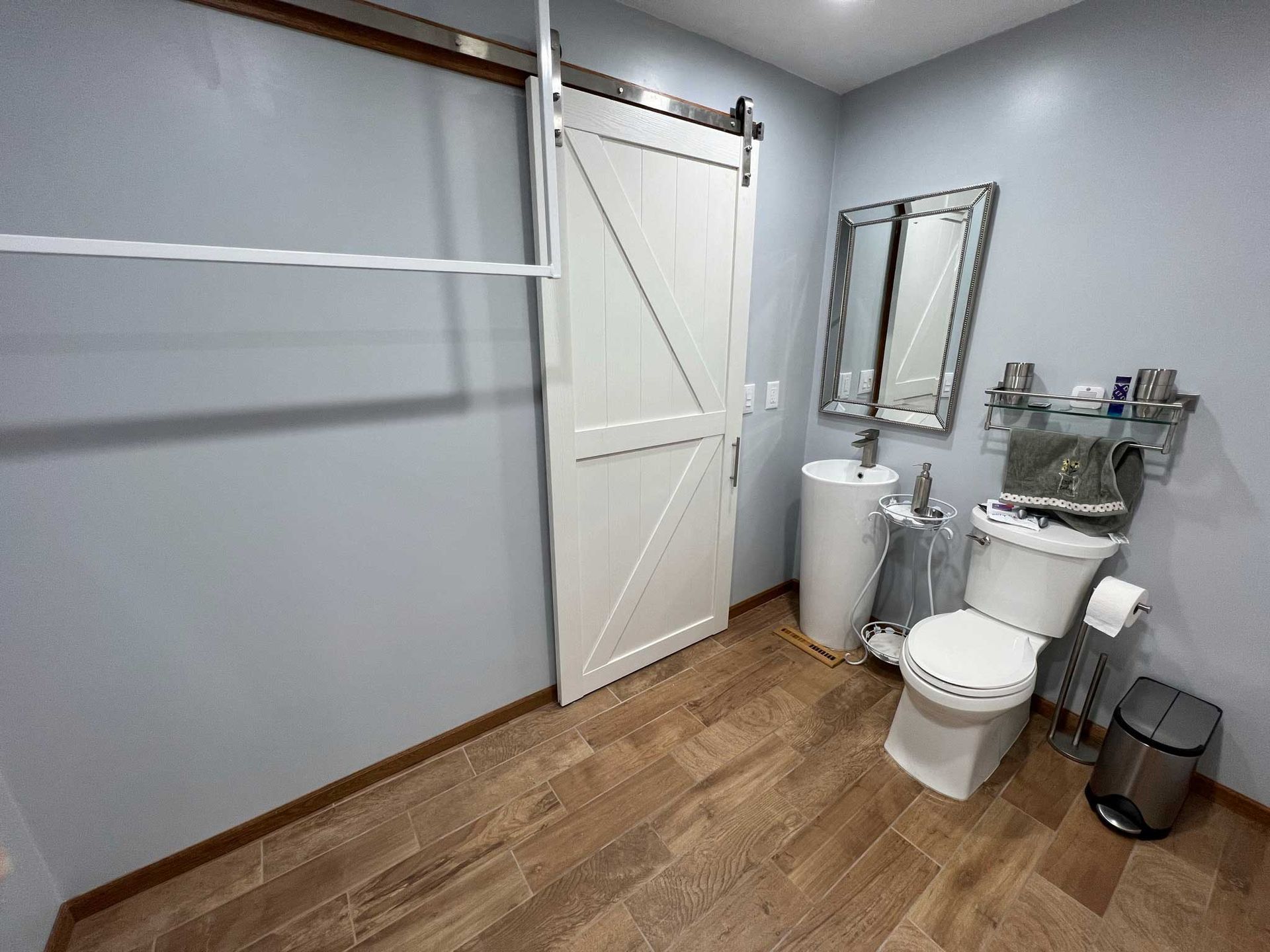 Bathroom with white sliding door, toilet, sink, mirror, and wood-look tile floor. Blue-gray walls.