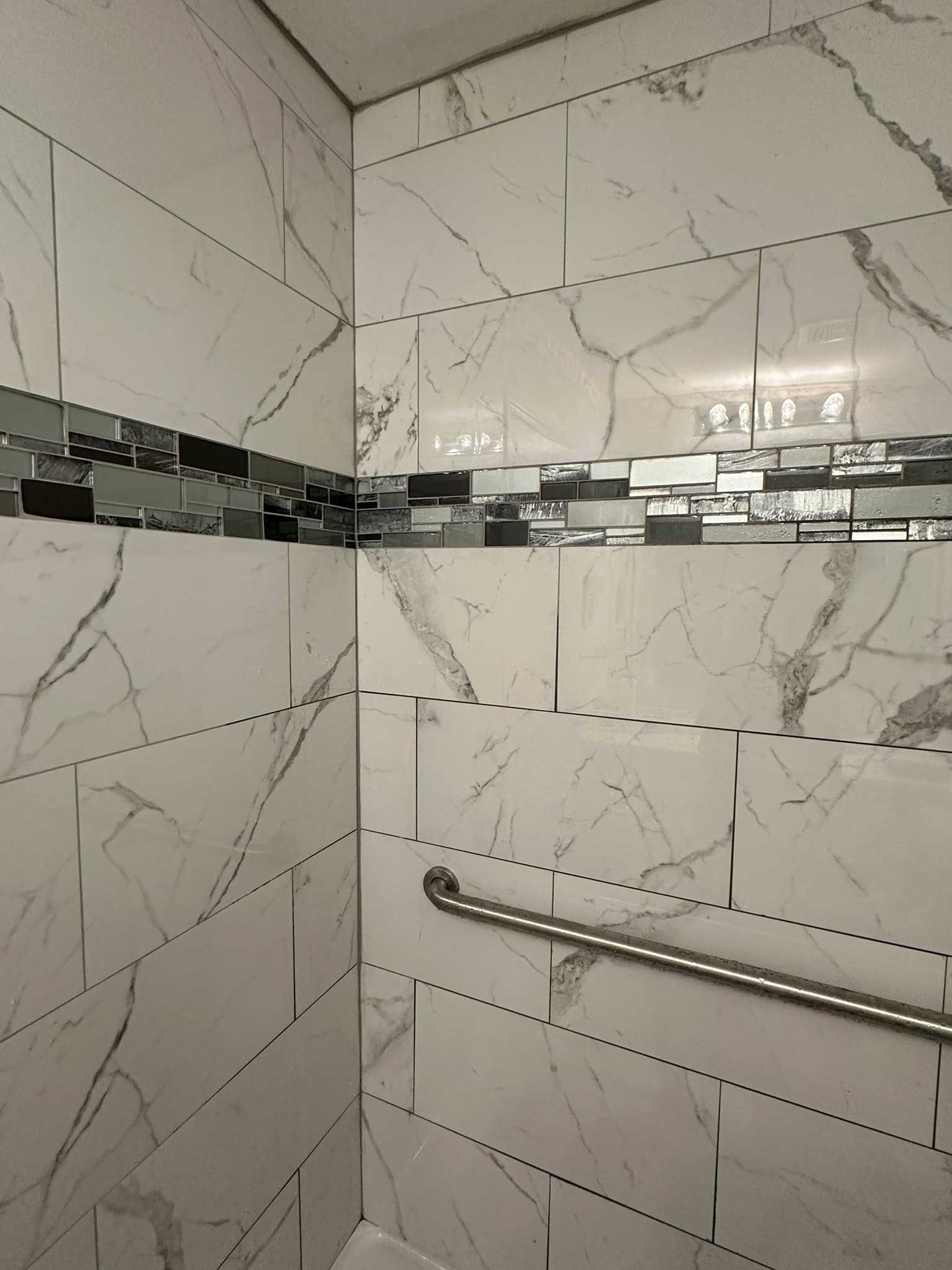White tiled shower with gray veining and decorative strip. A silver handrail is mounted on the wall.
