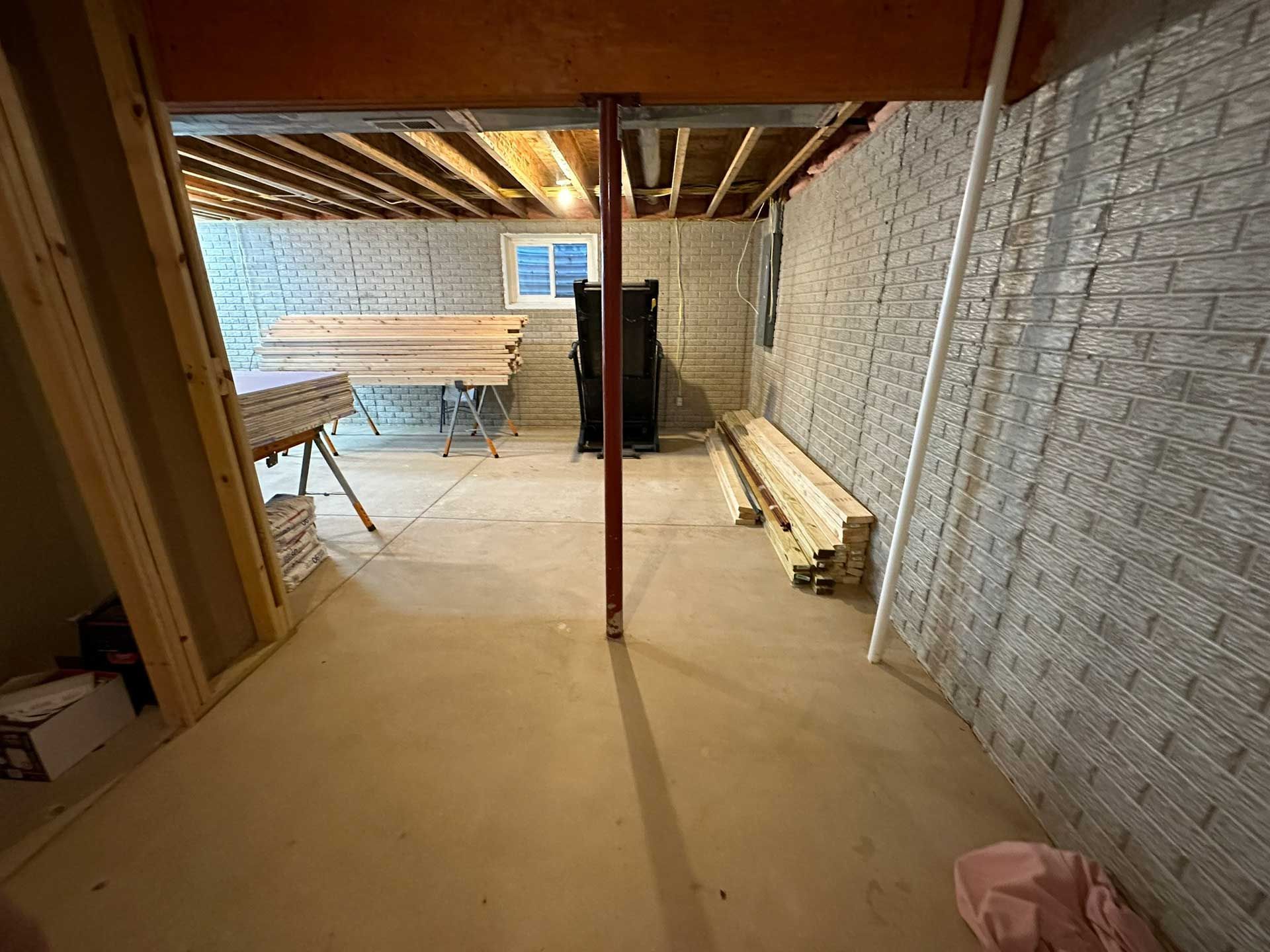Basement with concrete floor, brick-patterned walls, wooden beams, a support post, and construction materials.