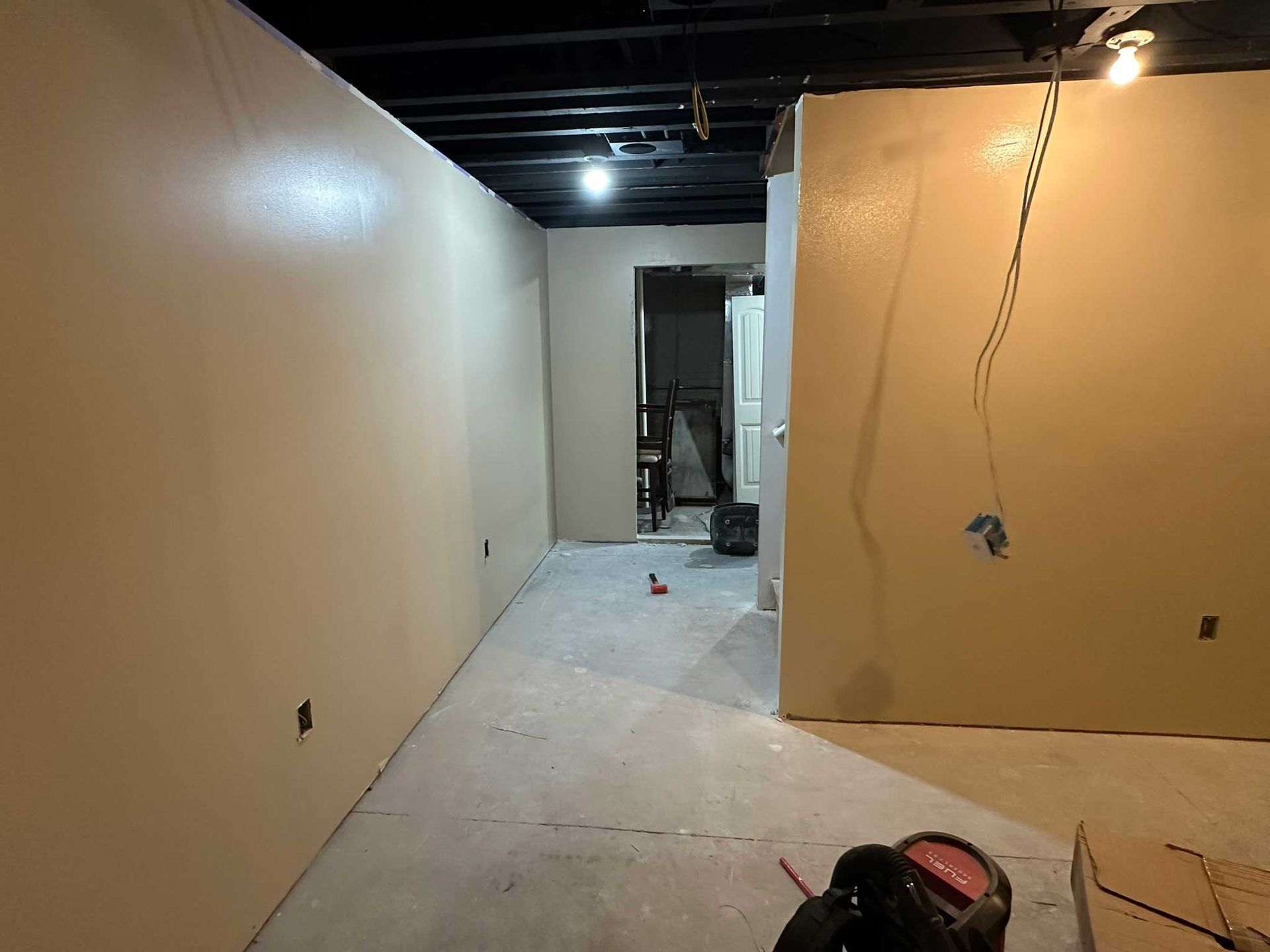 Bare-bones basement with beige walls, exposed wiring, black ceiling, and doorway to another room.
