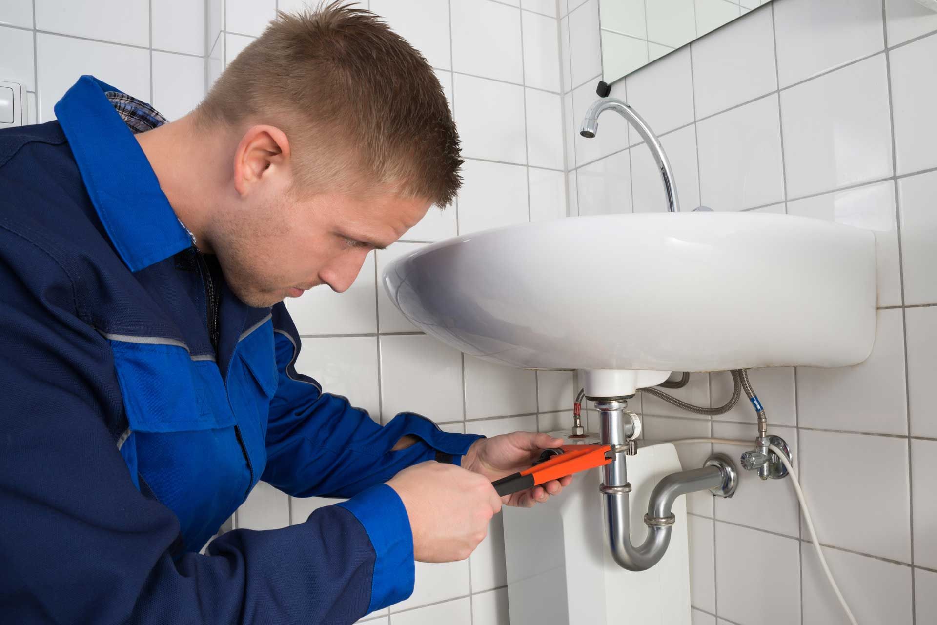 Plumber in blue overalls uses a wrench to fix a bathroom sink with white tiles in the background.