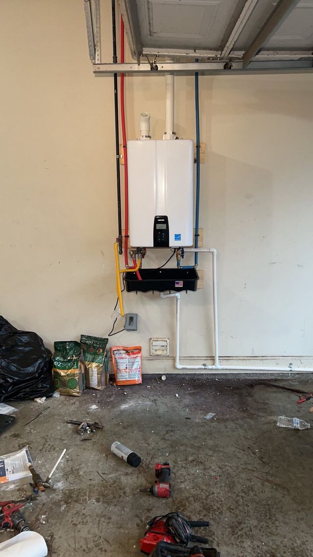 Gas water heater mounted on a garage wall. Piping and electrical components visible. Debris covers the floor.