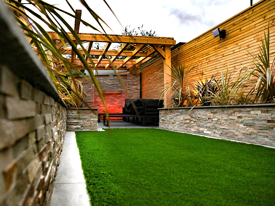 Lush green backyard with artificial turf, stone walls, wooden pergola, and a backdrop of wooden lattice and plantings.