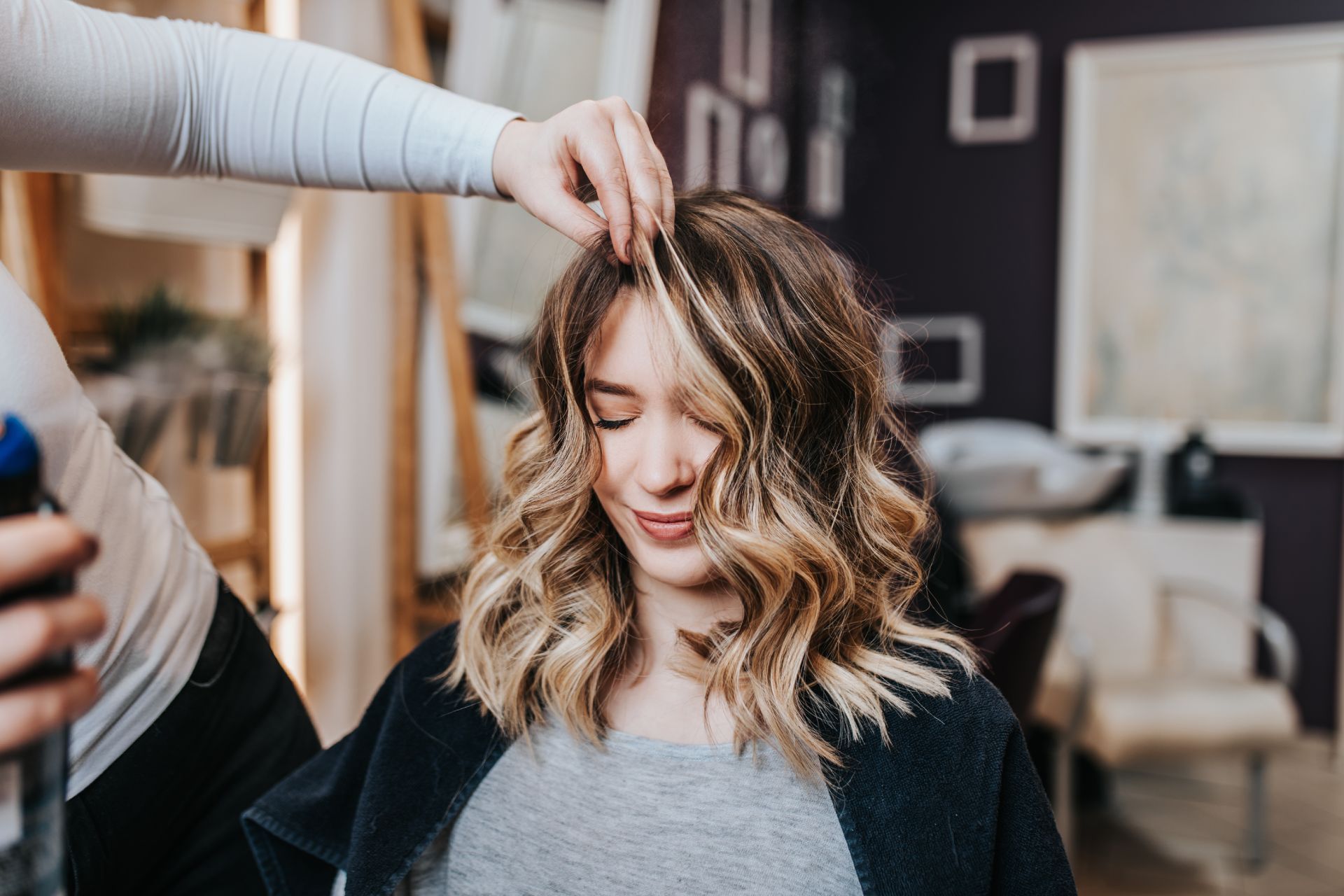 Hair stylist styling a client's wavy, highlighted hair at a salon. The client is smiling with her eyes closed.