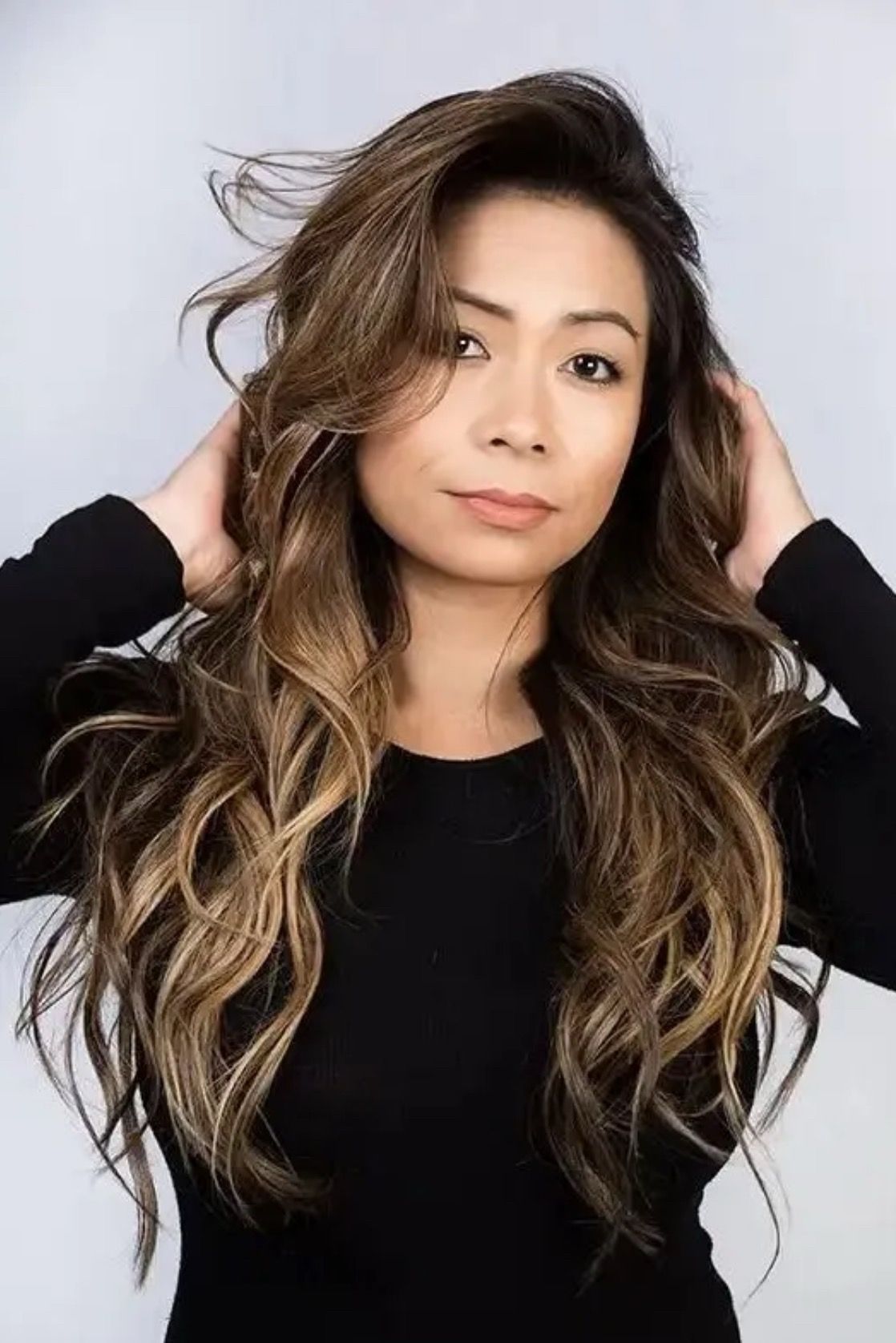 Woman with long, wavy brown hair, hands in hair, wearing black top, neutral expression.