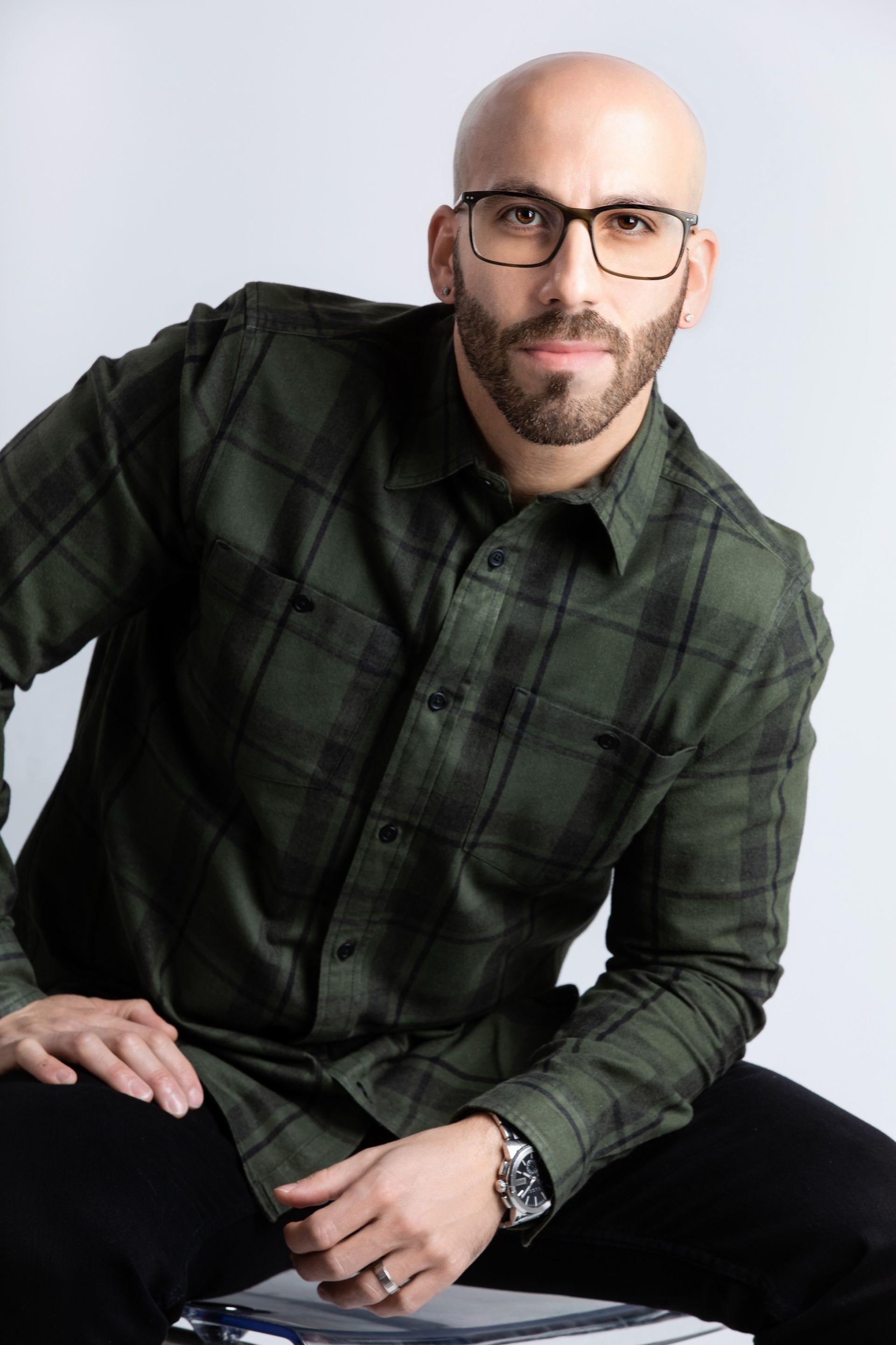 Man with glasses and a beard in a green plaid shirt, seated and looking towards the camera.