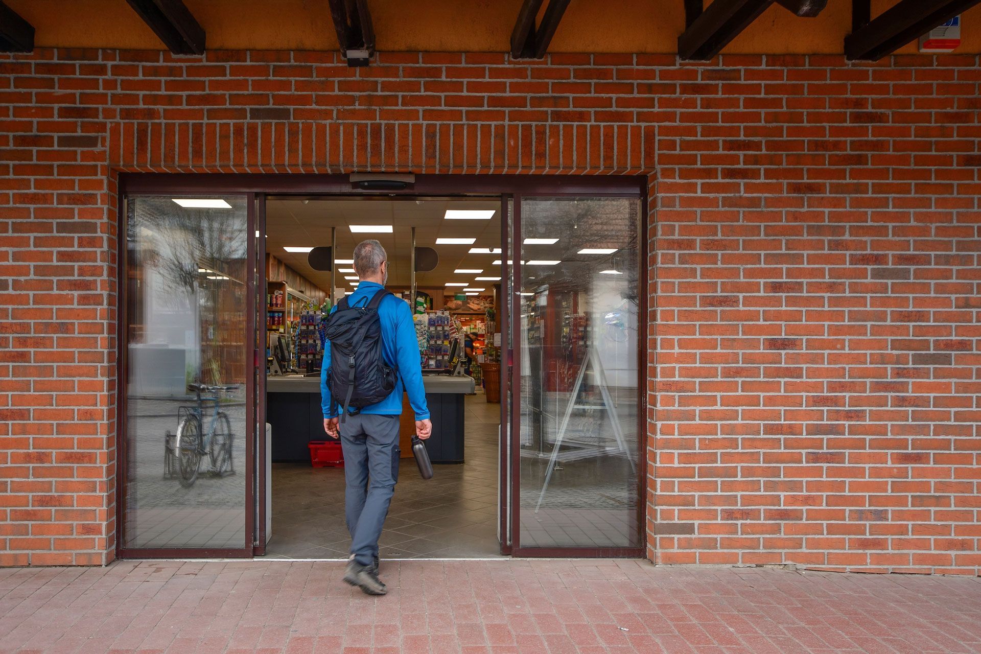 Person entering a store through automatic glass doors; brick exterior, shelves and goods visible inside.