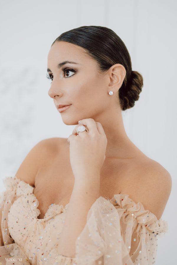 A woman wearing a wedding dress and earrings is touching her face.