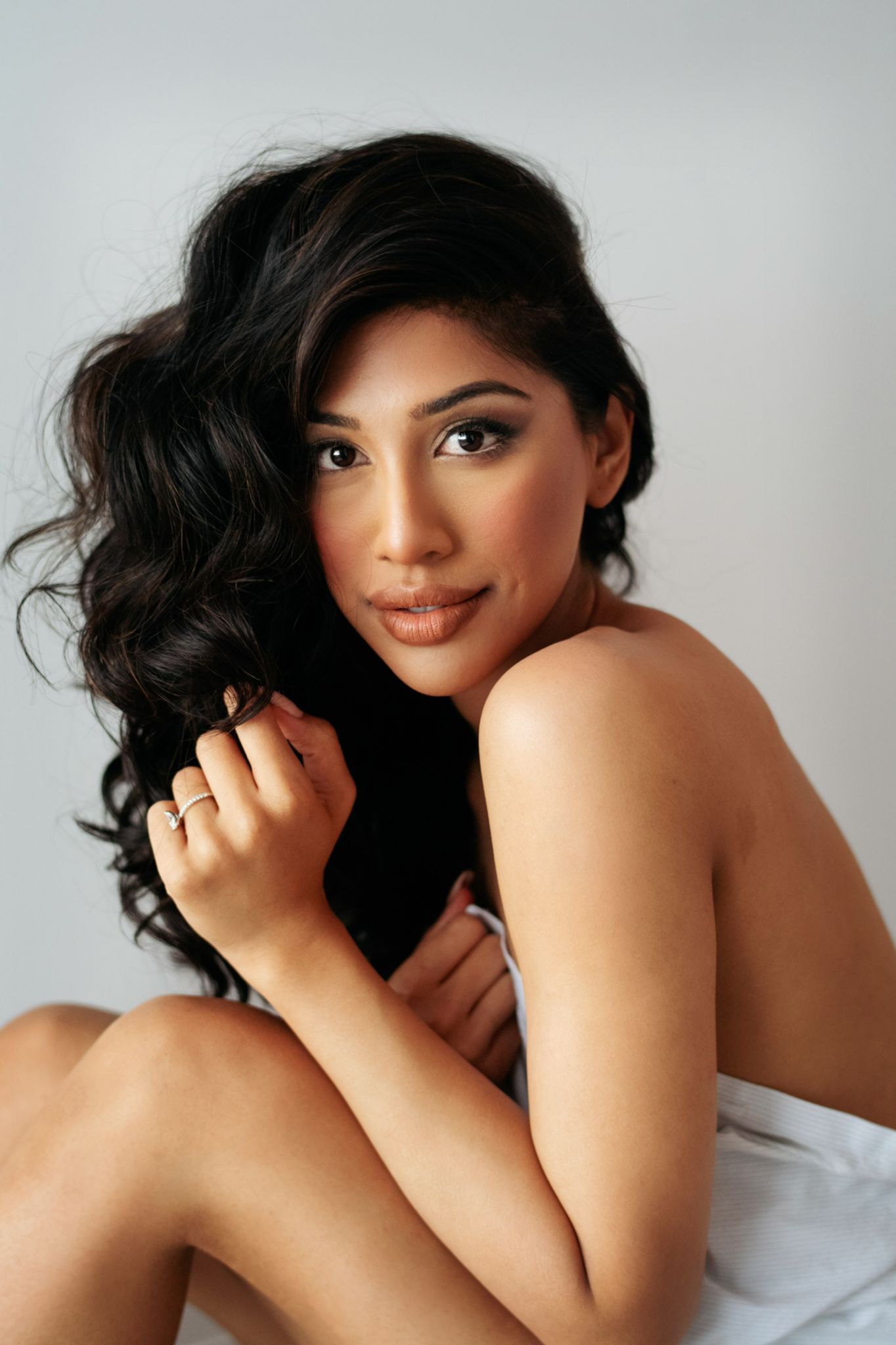 A woman without a shirt is sitting on a bed holding her hair.