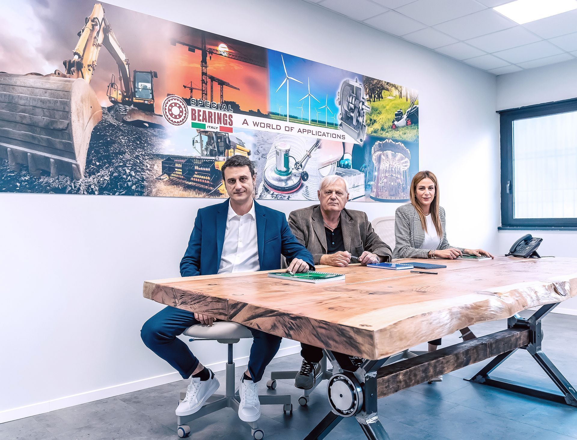 Tre persone sono sedute a un grande tavolo di legno in un ufficio. Sulla parete è appeso un murale raffigurante attrezzature edili e energie rinnovabili.