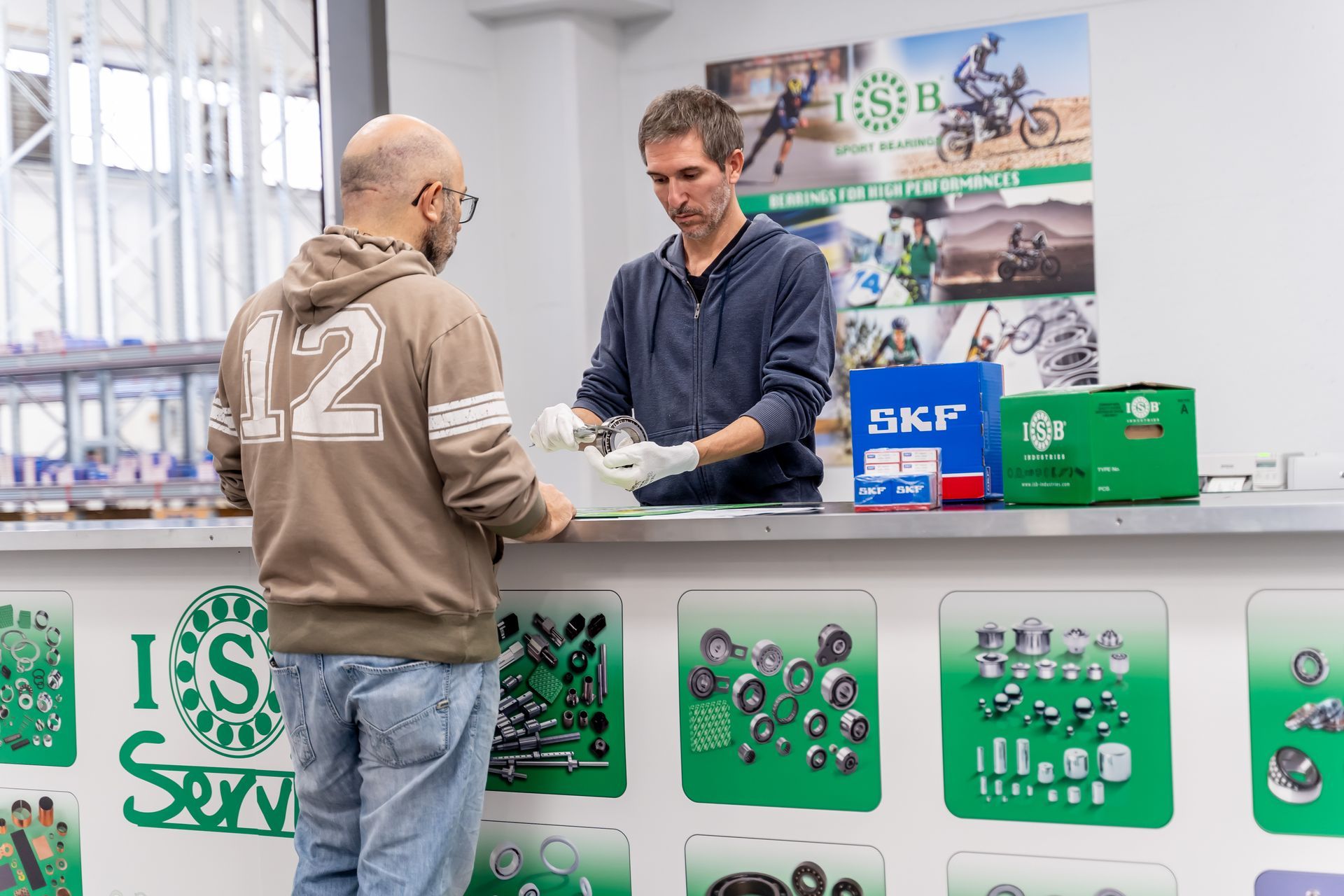 Uomo che acquista pezzi di ricambio al bancone di un negozio, il commesso maneggia l'articolo. Interno del negozio, pezzi esposti.