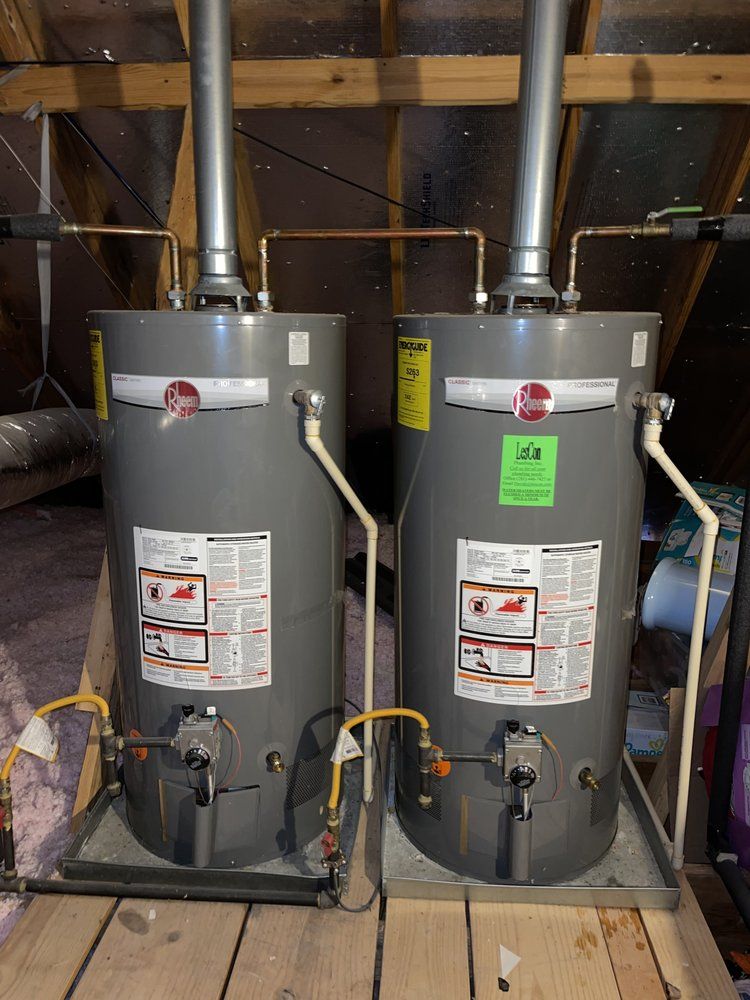 Two water heaters are sitting on a wooden floor in an attic.