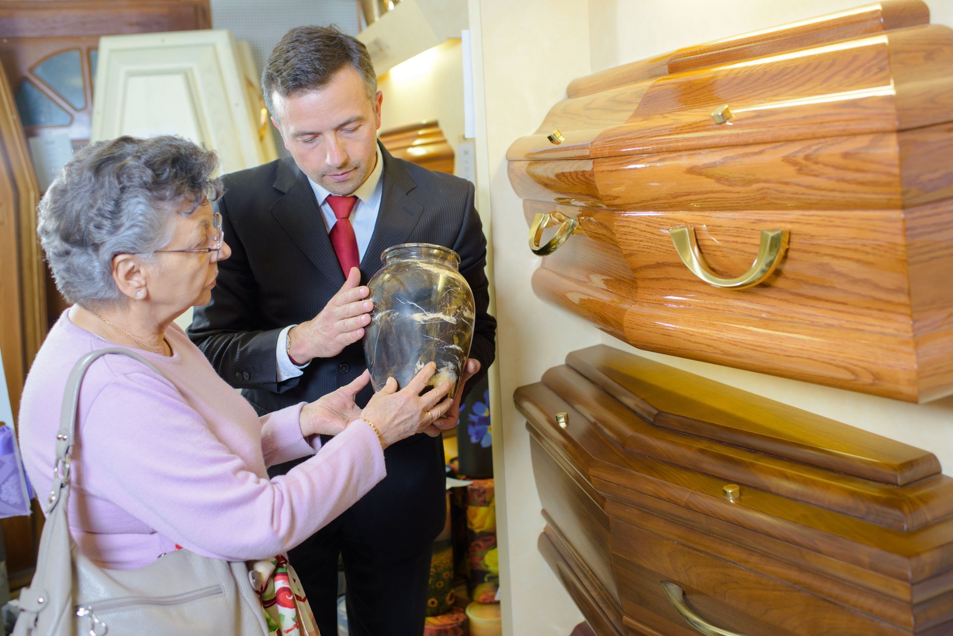 A senior woman looking at an urn being held by a funeral director next to coffins on display. A senior woman looking at an urn being held by a funeral director next to coffins on display.