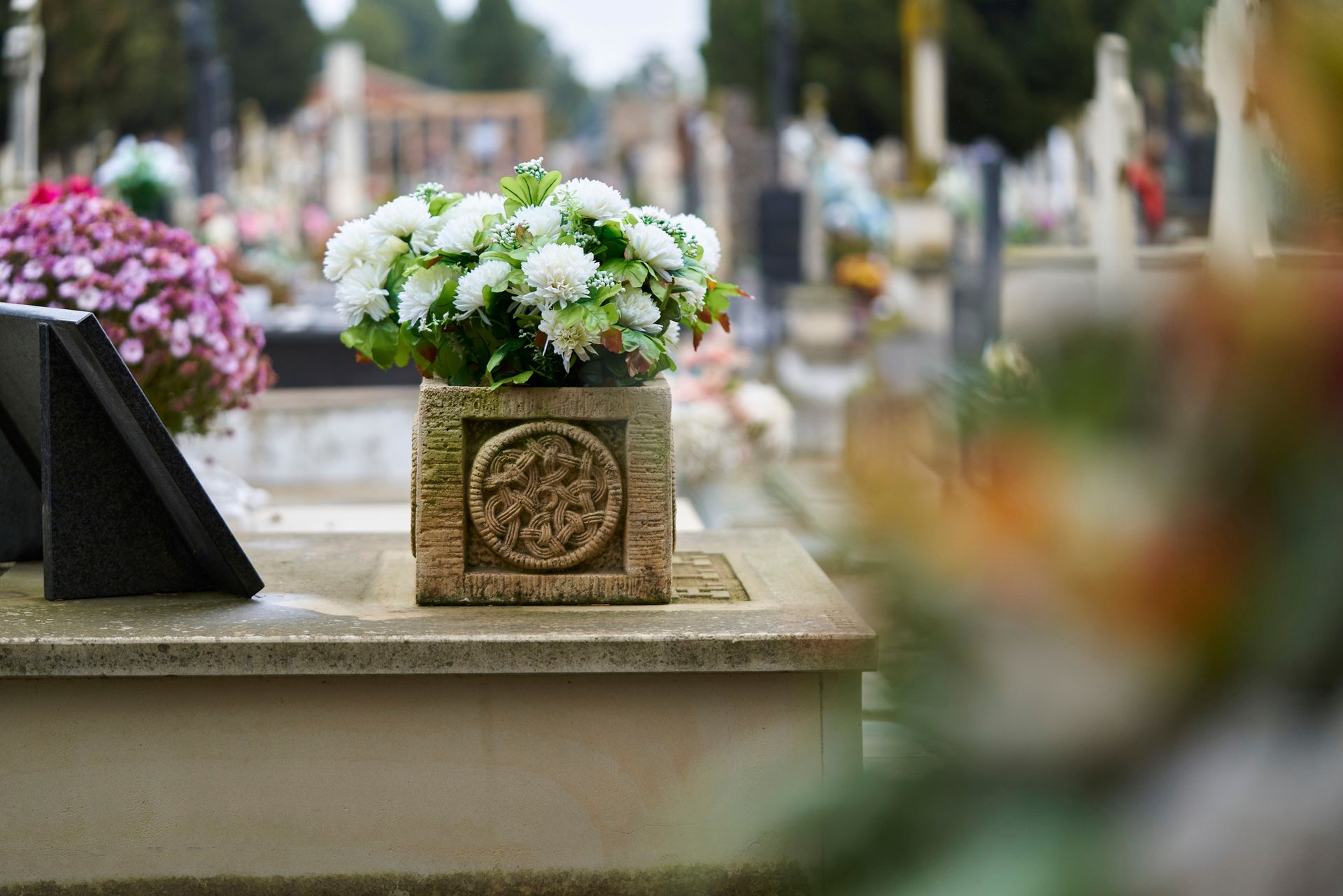 A stone flowerpot with white chrysanthemums placed on a grave in a cemetery surrounded by headstones