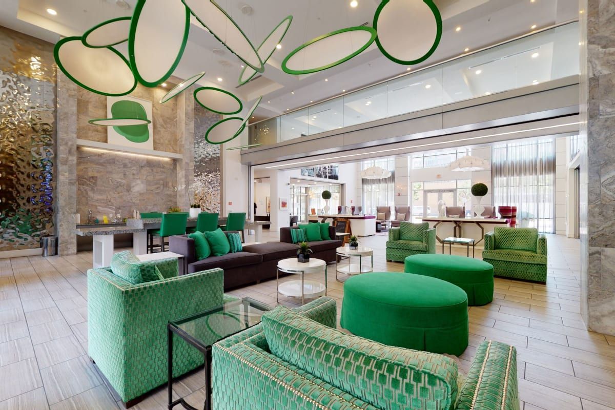 There are many green chairs and ottomans in the lobby of a hotel.