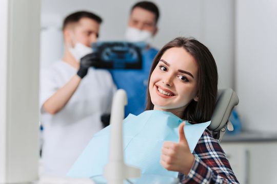 Smiling dental patient giving thumbs up in a clinic, with dentists blurred in the background