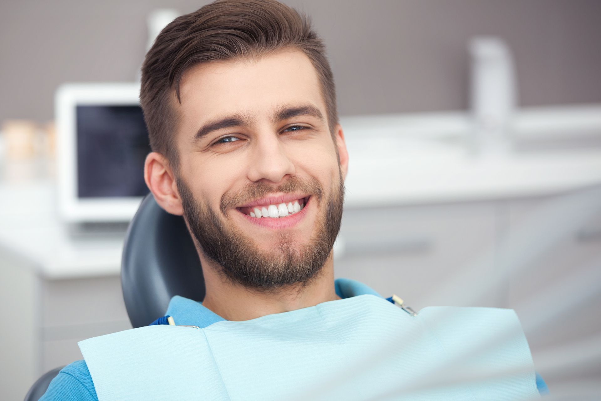Smiling patient in a dental chair wearing a blue bib, with a blurred clinic background