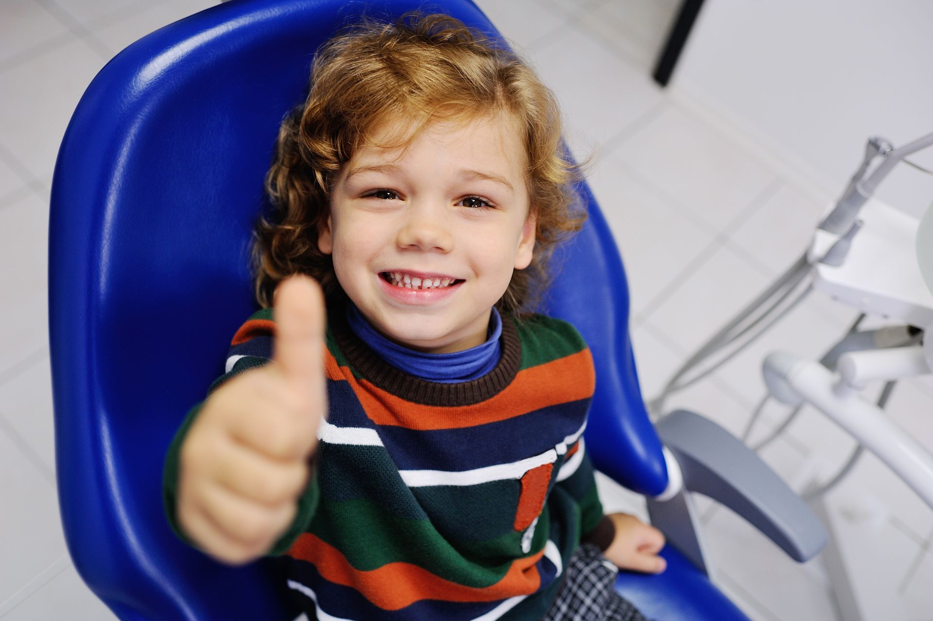 Smiling child in a blue chair giving a thumbs-up in a dental office