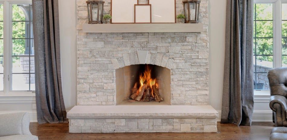 A lit fireplace in a stone brick wall with framed artwork, flanked by windows with curtains.