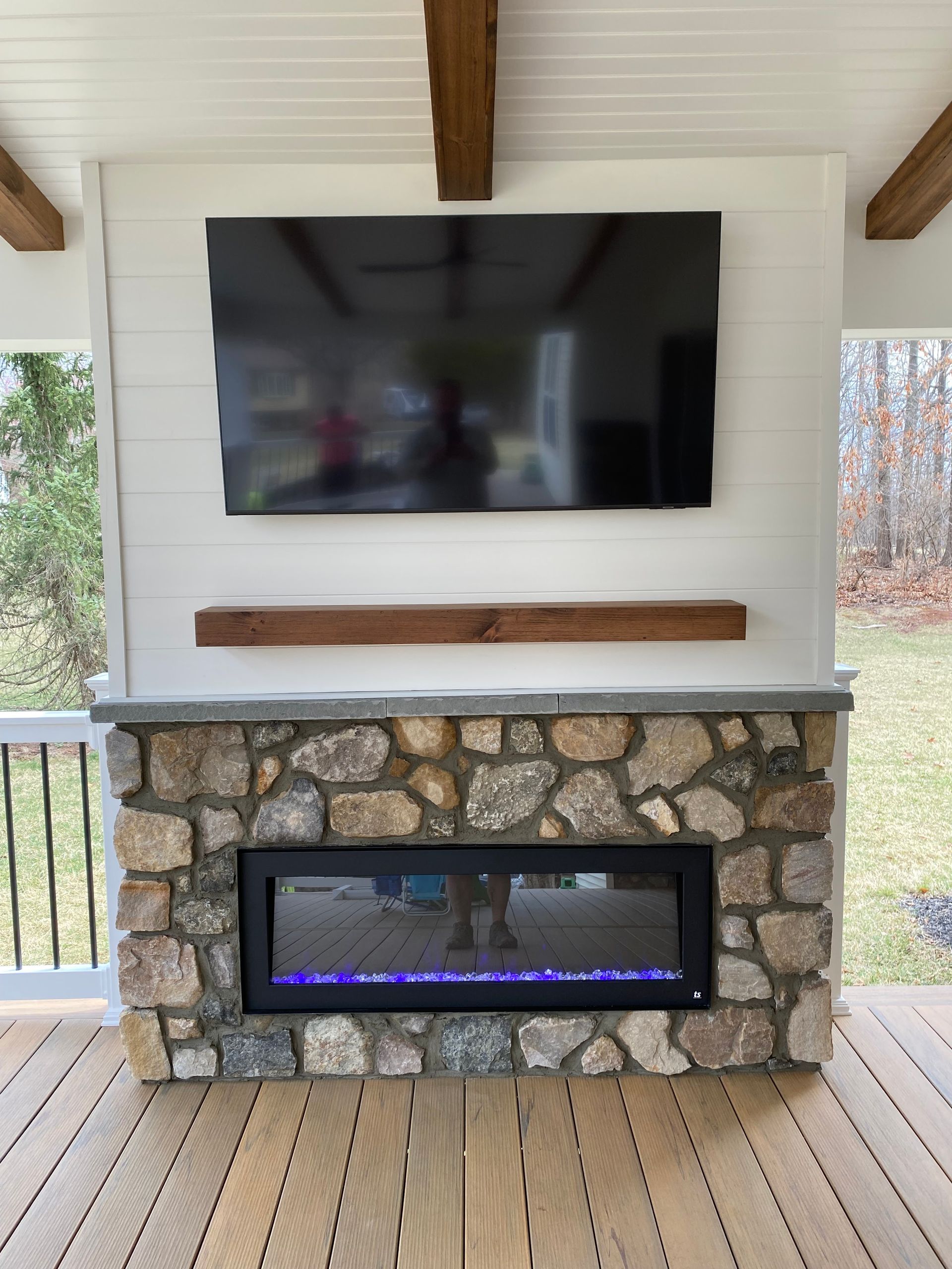 Stone fireplace with mounted TV on a white shiplap wall, wooden mantel, and beams, on a wooden deck.