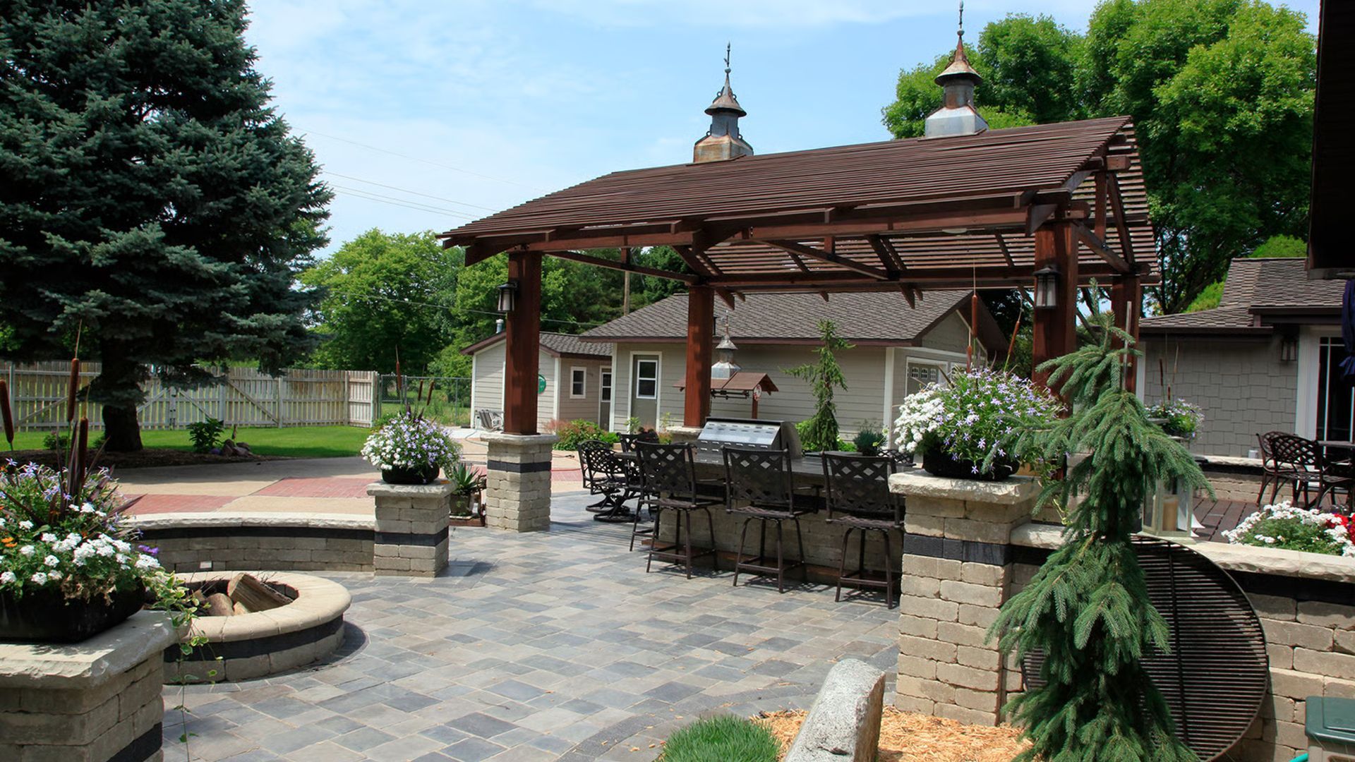 Backyard patio with pergola, grill, seating, and landscaping; a building with two towers in the background.