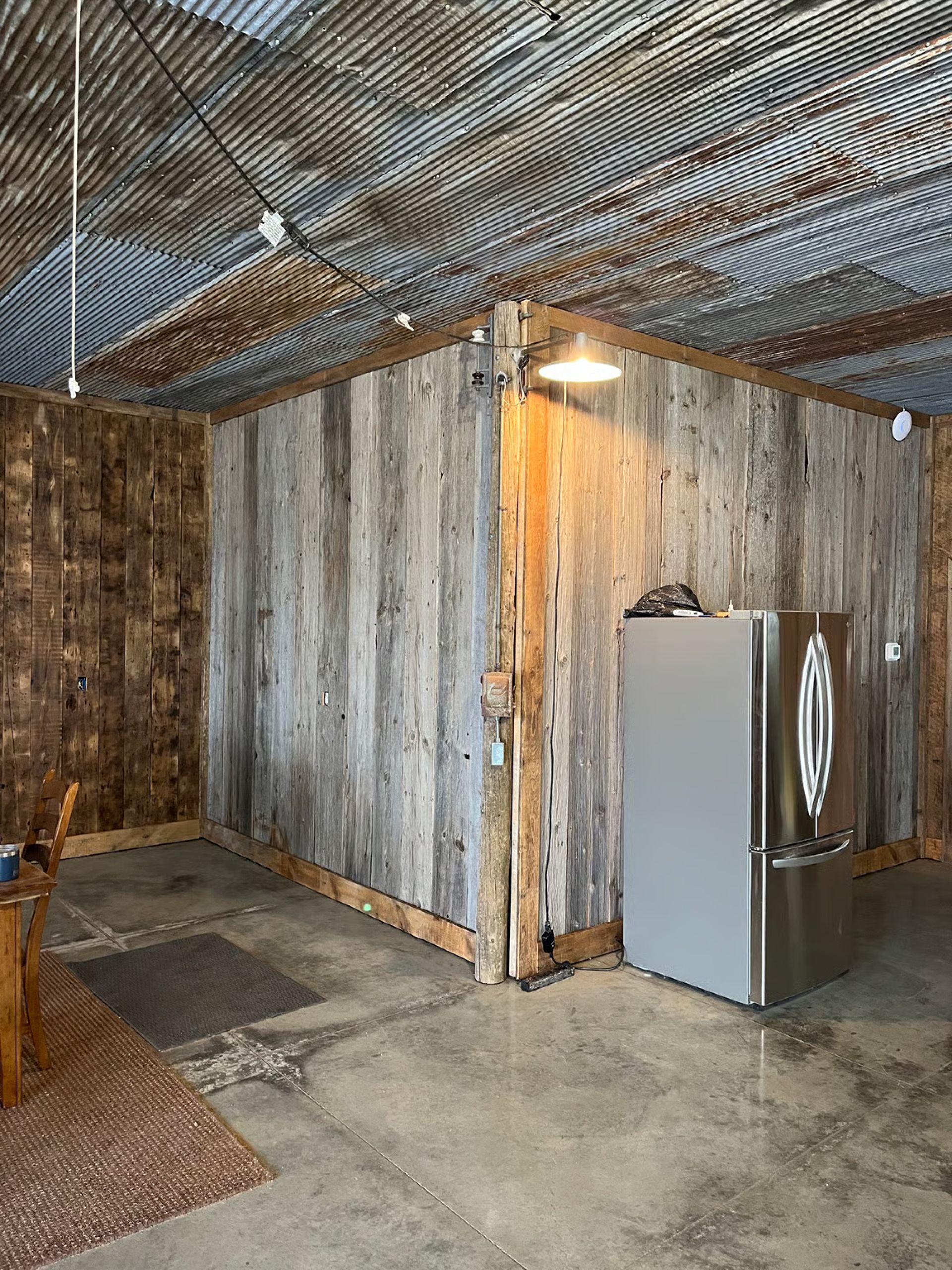 Rustic kitchen with weathered wood walls, corrugated metal ceiling, concrete floor, stainless steel refrigerator.