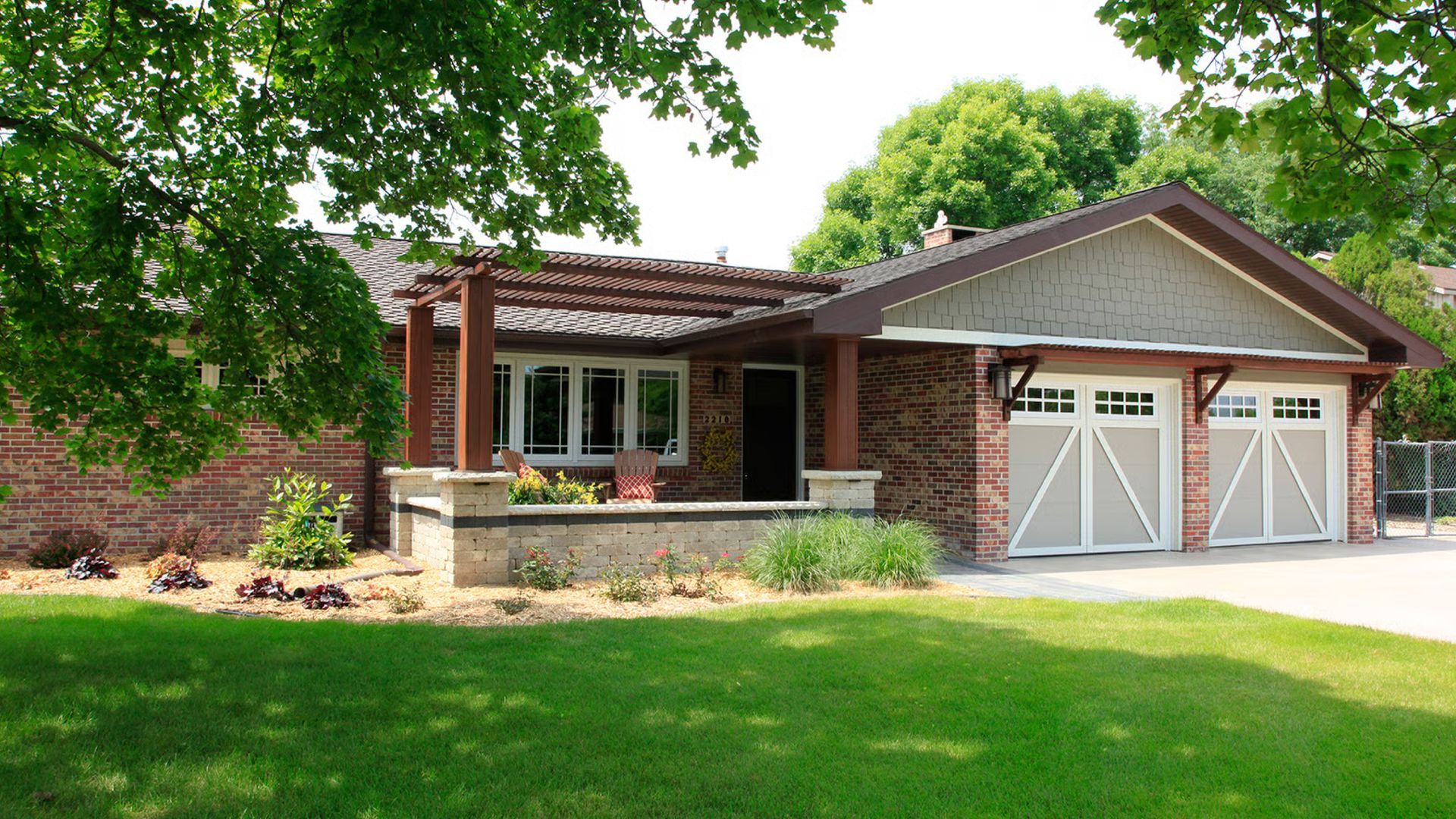 Brick house with attached garage, pergola, and green lawn. White garage doors, brown roof.