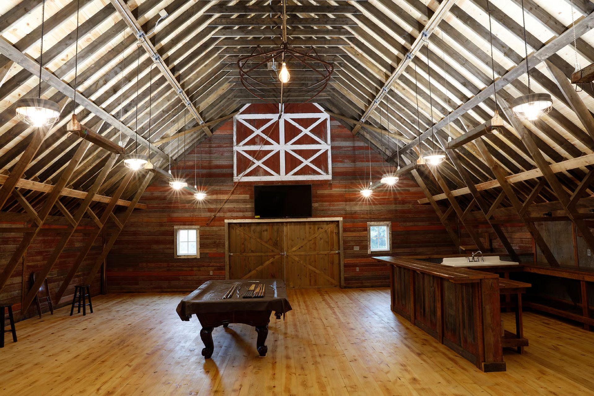 Interior of a rustic barn with wooden beams, a bar, pool table, and hanging lights.