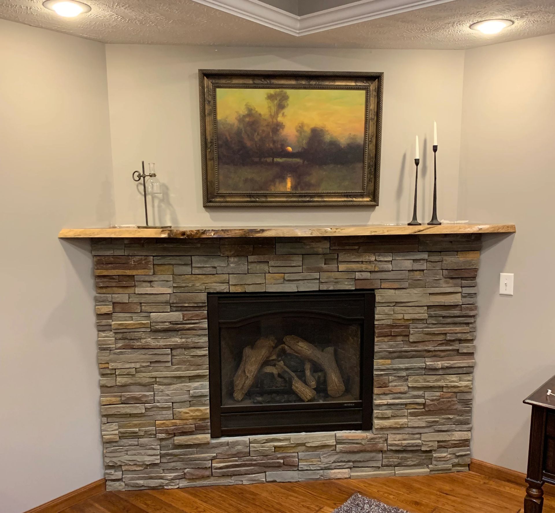 Fireplace with stone facade, wooden mantel, and artwork above in a corner setting.