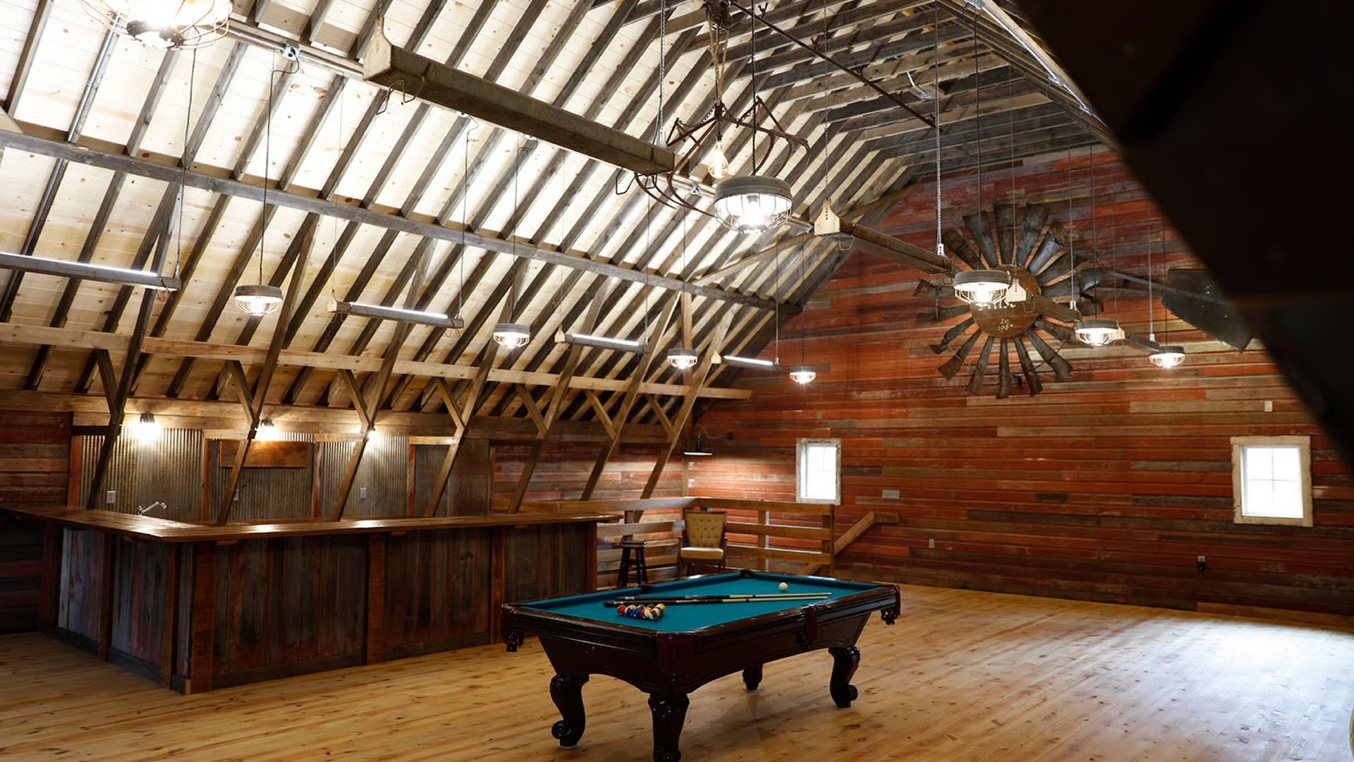 Rustic barn interior with a pool table, bar, and wooden ceiling.