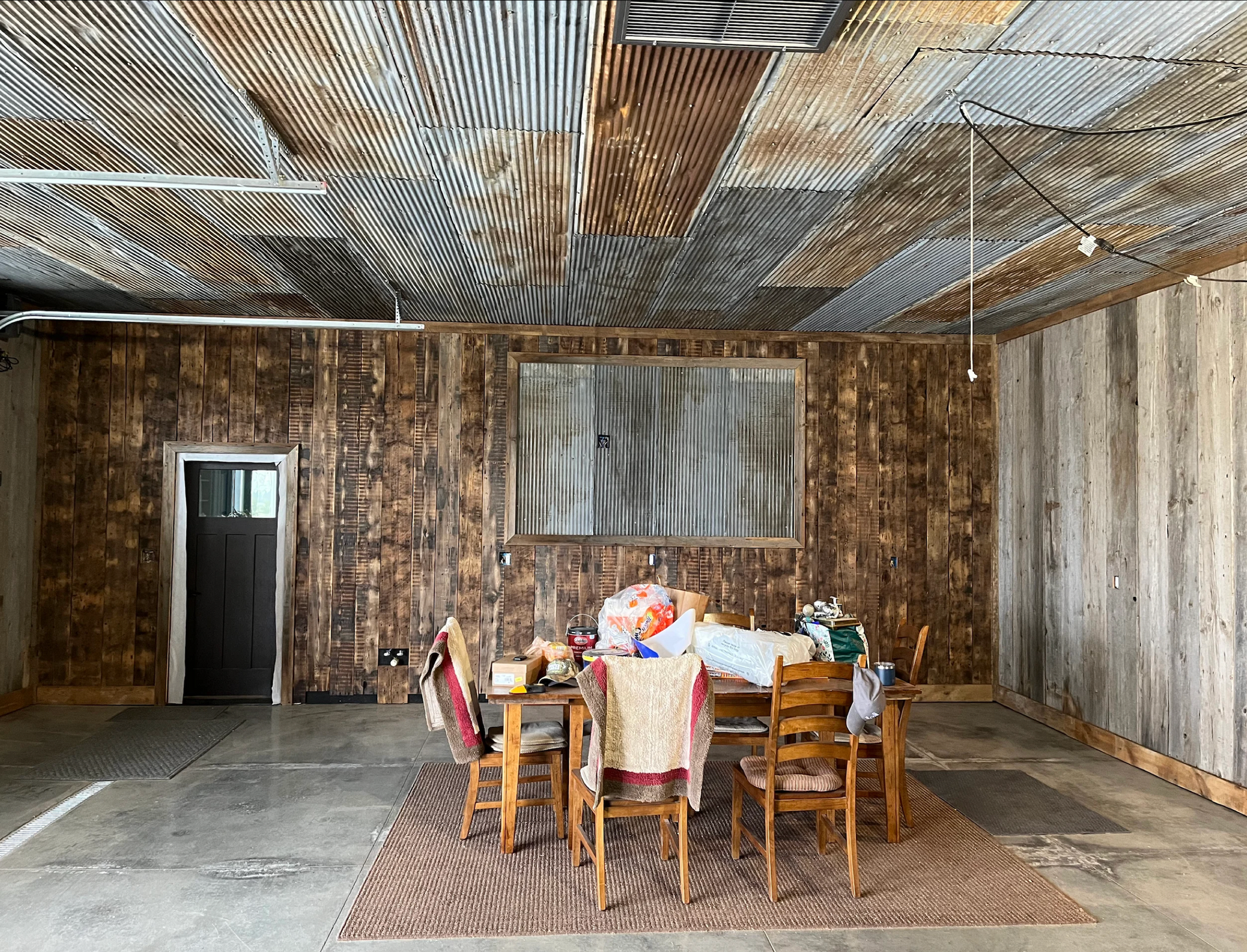 Rustic dining area with corrugated metal ceiling and wood-paneled walls. Table set with chairs, rug on concrete floor.