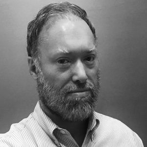 Man with beard and thinning hair, wearing a striped shirt, looking towards the camera.