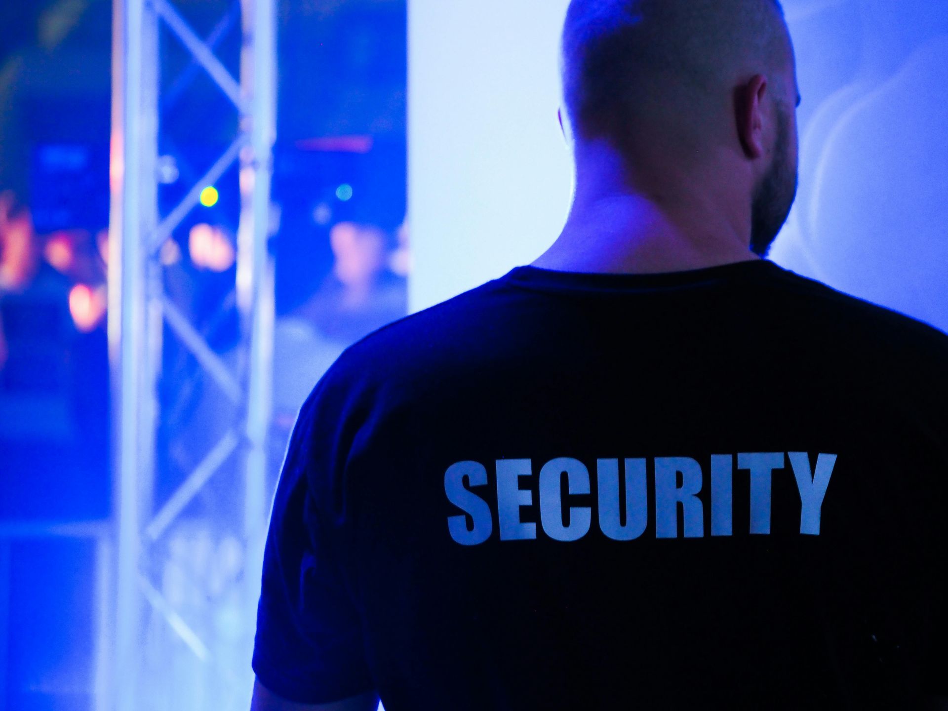 A man wearing a security shirt is standing in front of a blue light.