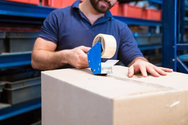 A man is using a tape dispenser to tape a box in a warehouse. - Port Saint John, FL - Shipping Depot of Port St John