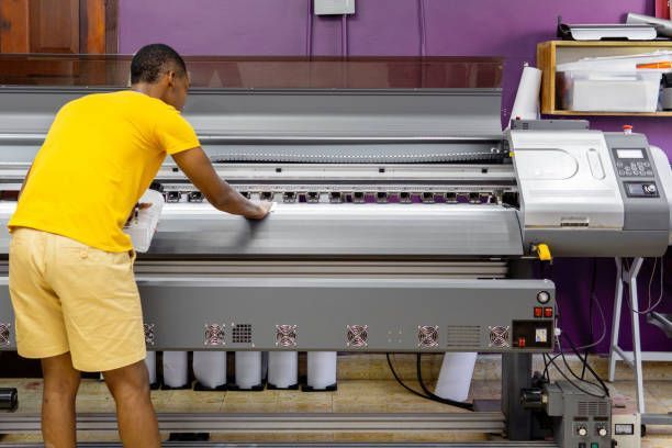 A man in a yellow shirt is standing in front of a large printer. - Port Saint John, FL - Shipping Depot of Port St John