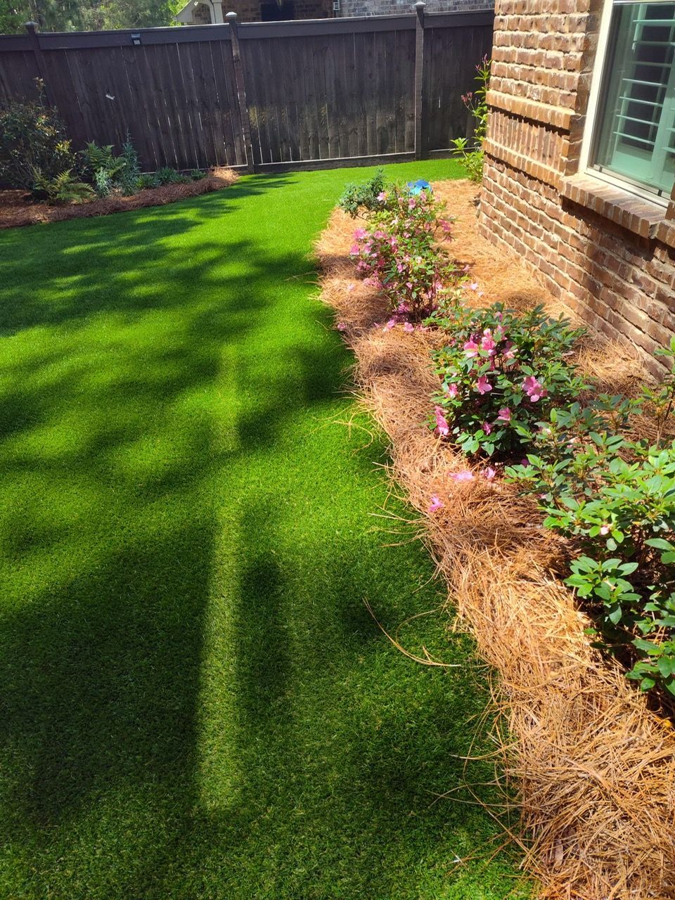 A lush green lawn next to a brick house with flowers and a fence.