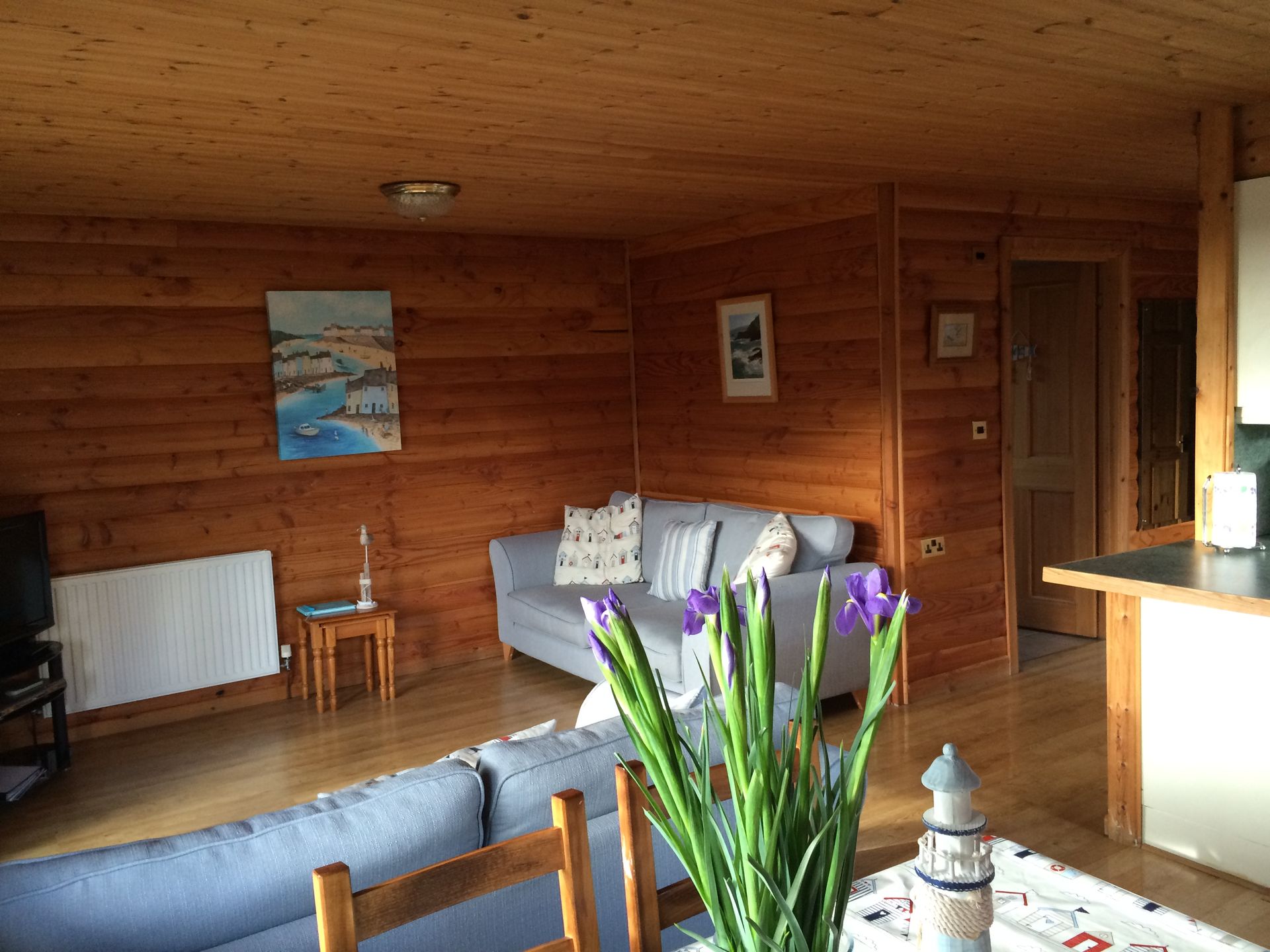 A cozy living room with light wooden log walls, a gray sofa, a white radiator, and blue flowers in the foreground.