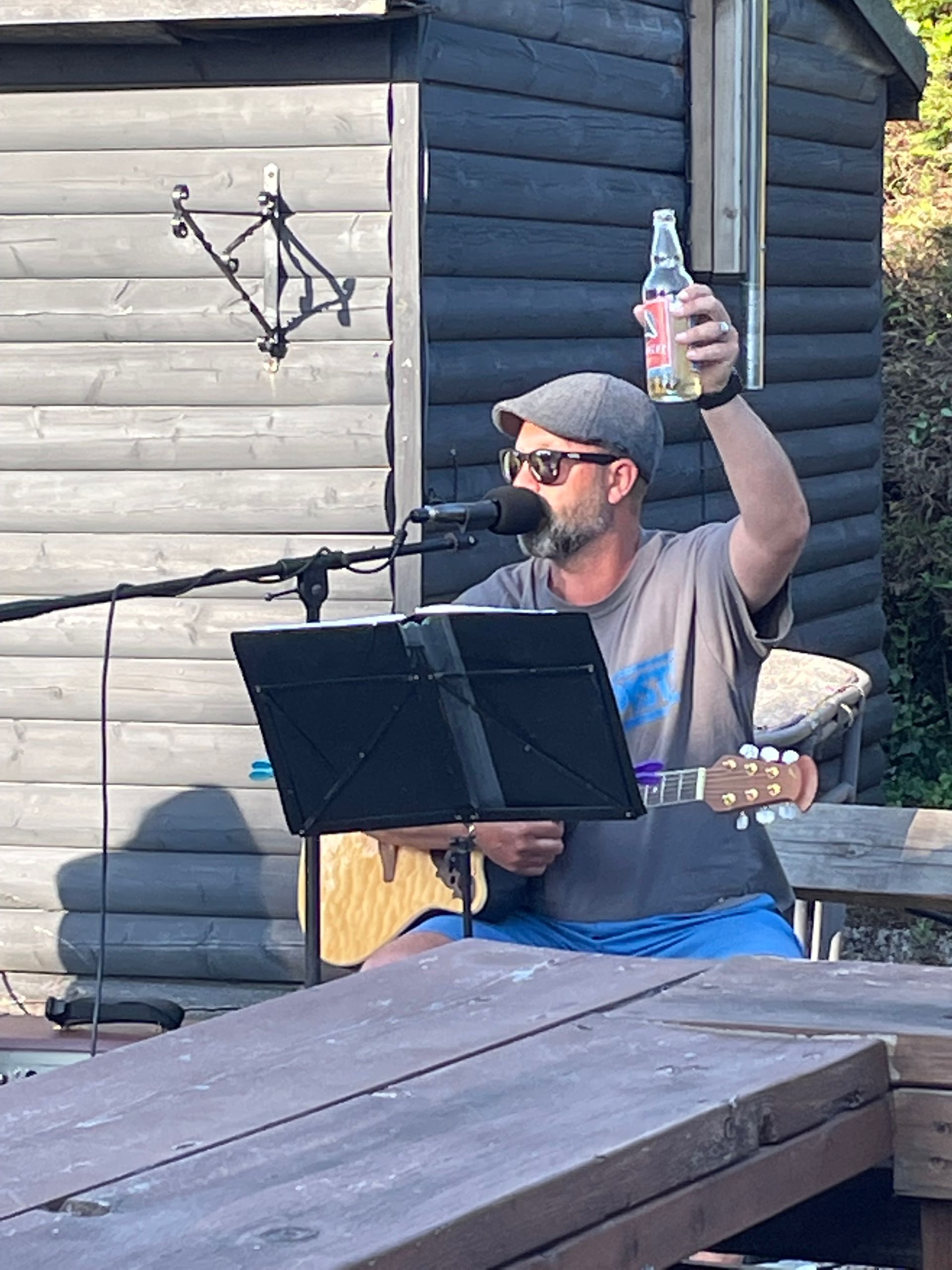 A person wearing a cap and sunglasses plays guitar and sings into a microphone, raising a clear bottle outdoors.