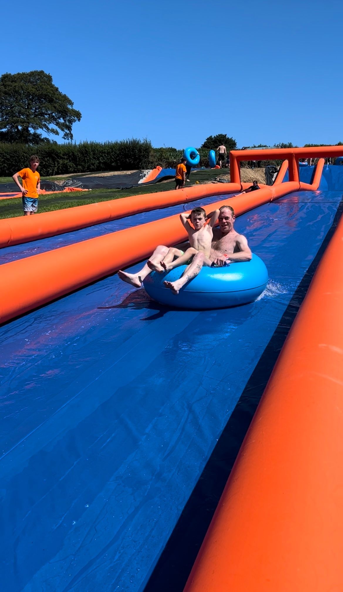 An adult and child ride a blue tube down a long, blue inflatable water slide with orange side bumpers under a clear sky.