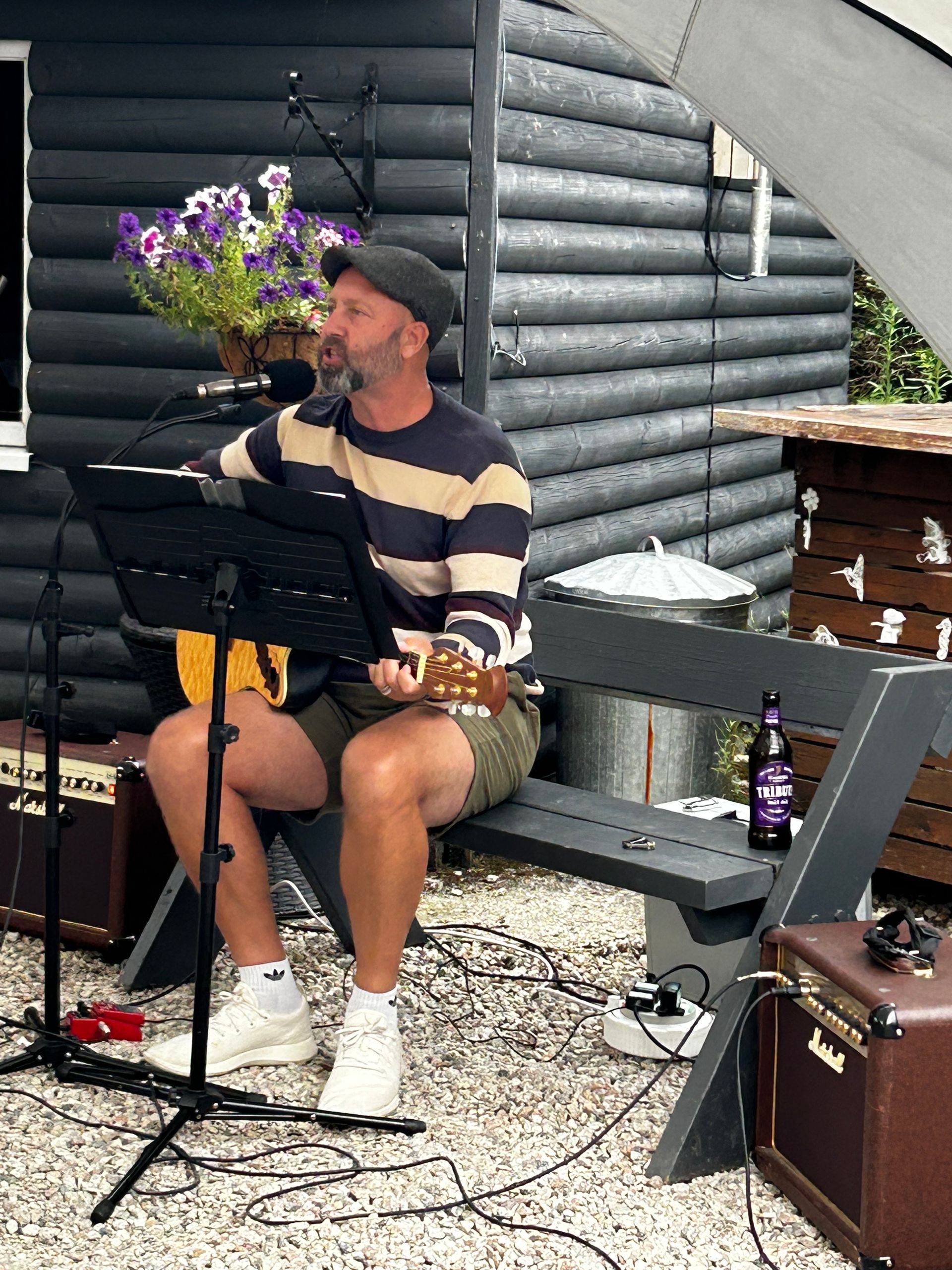 A bearded musician in a striped sweater and cap plays guitar and sings into a microphone while sitting on a garden bench.