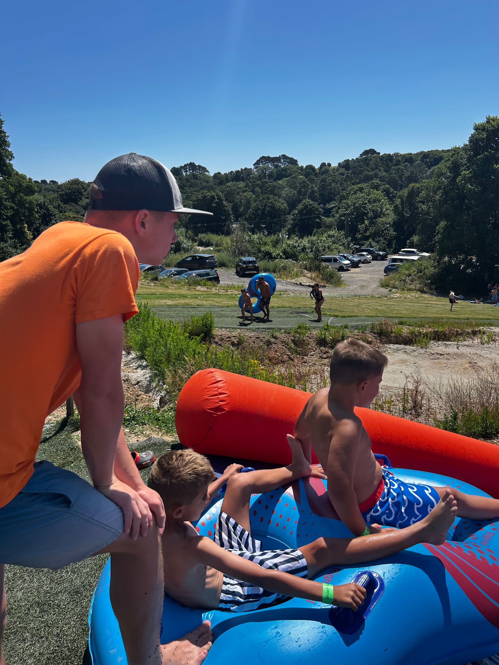 A man in an orange shirt and two children sit on a blue inflatable tube on a sunny day at an outdoor park.