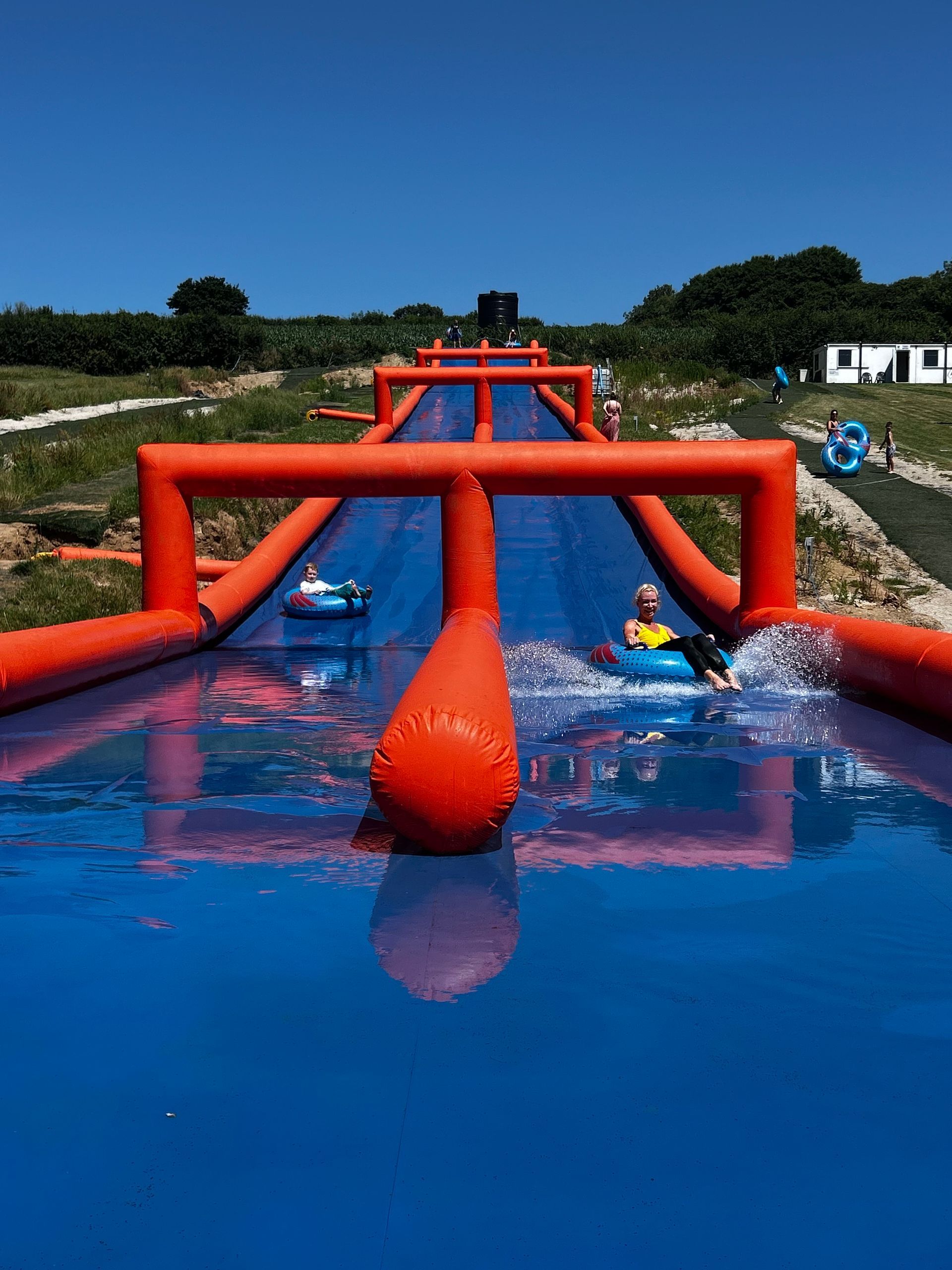 People ride inflatable tubes down a long, blue water slide framed by bright orange, air-filled arches on a sunny day.