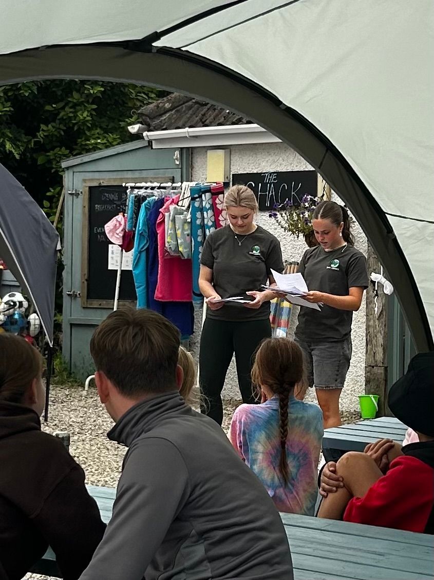 Two people in matching gray t-shirts stand outside under a canopy, speaking to a group while holding papers and clipboards.